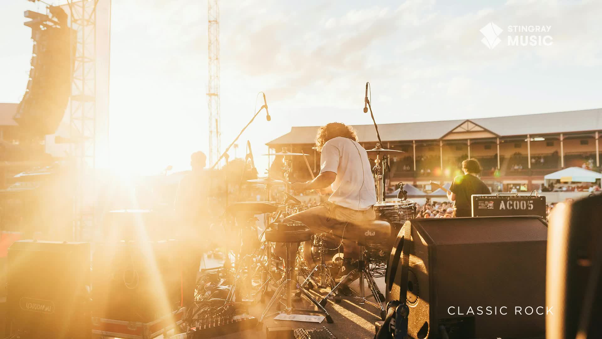The drummer, bathed in sunlight, is pounding away at his kit, the cymbals catching the golden light. The stage is set for a classic rock concert, with a crowd gathered in the distance.

