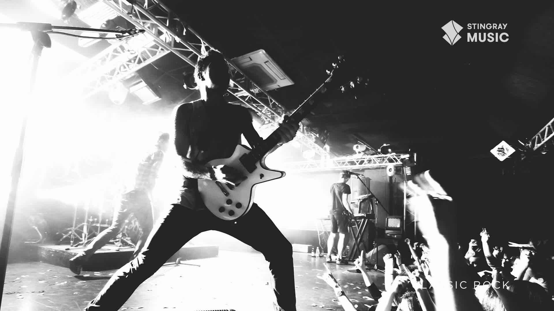The guitarist, backlit by stage lights, is mid-stride, leaning into his instrument. The crowd's hands are raised, reaching towards the stage as the band plays a set on Stingray Classic Rock.
