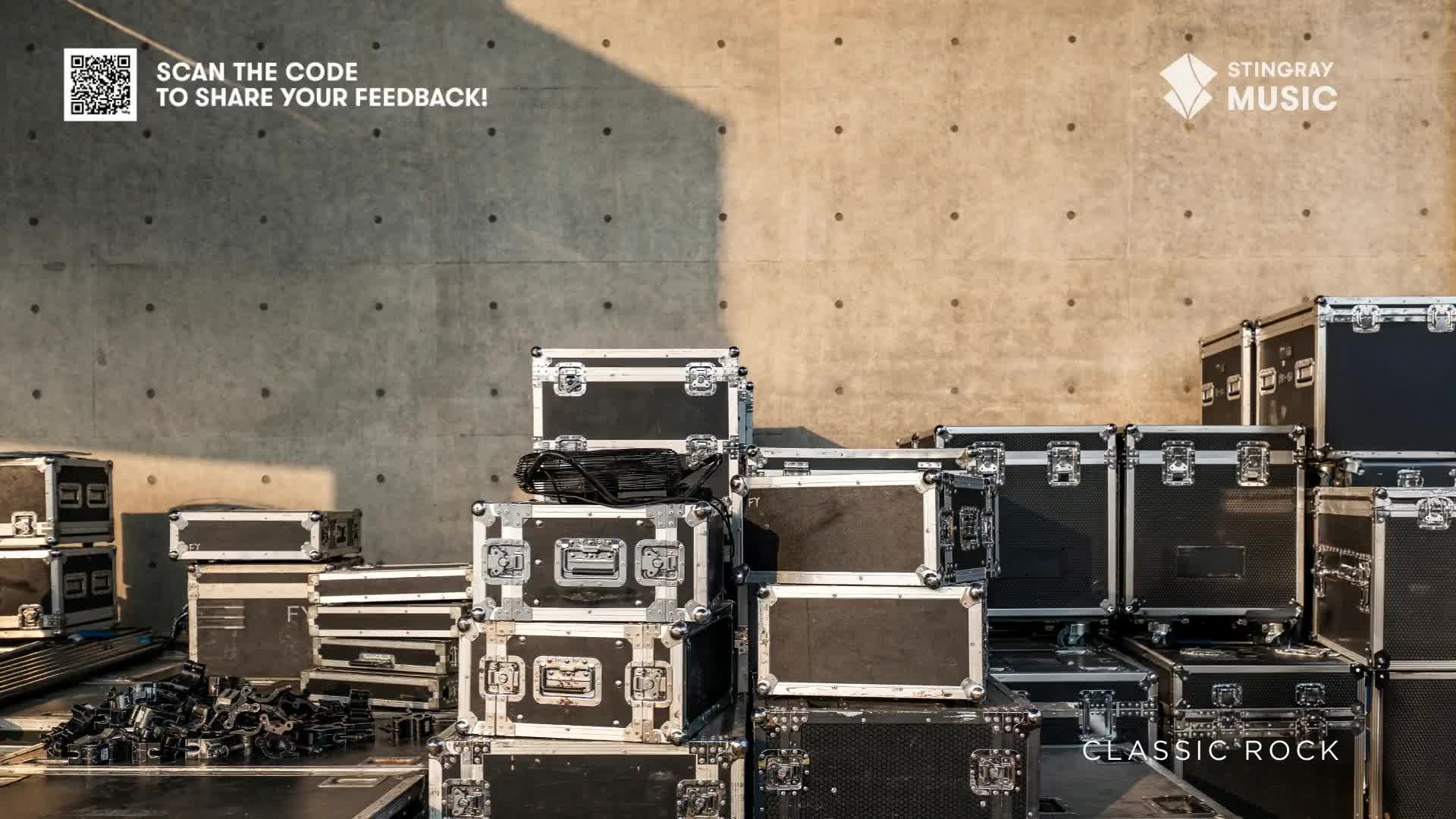 The gear cases are stacked high, ready for a Stingray Classic Rock show. Sunlight casts shadows across the concrete wall, illuminating the equipment.
