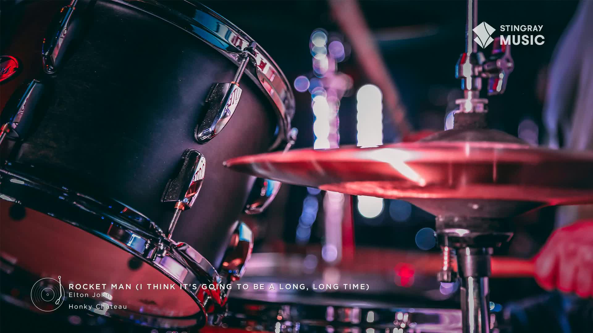 A drum kit, lit with red stage lights, fills the foreground. The drummer's stick is about to strike a cymbal as Elton John's "Rocket Man" blasts from the Stingray Classic Rock station in Canada.

