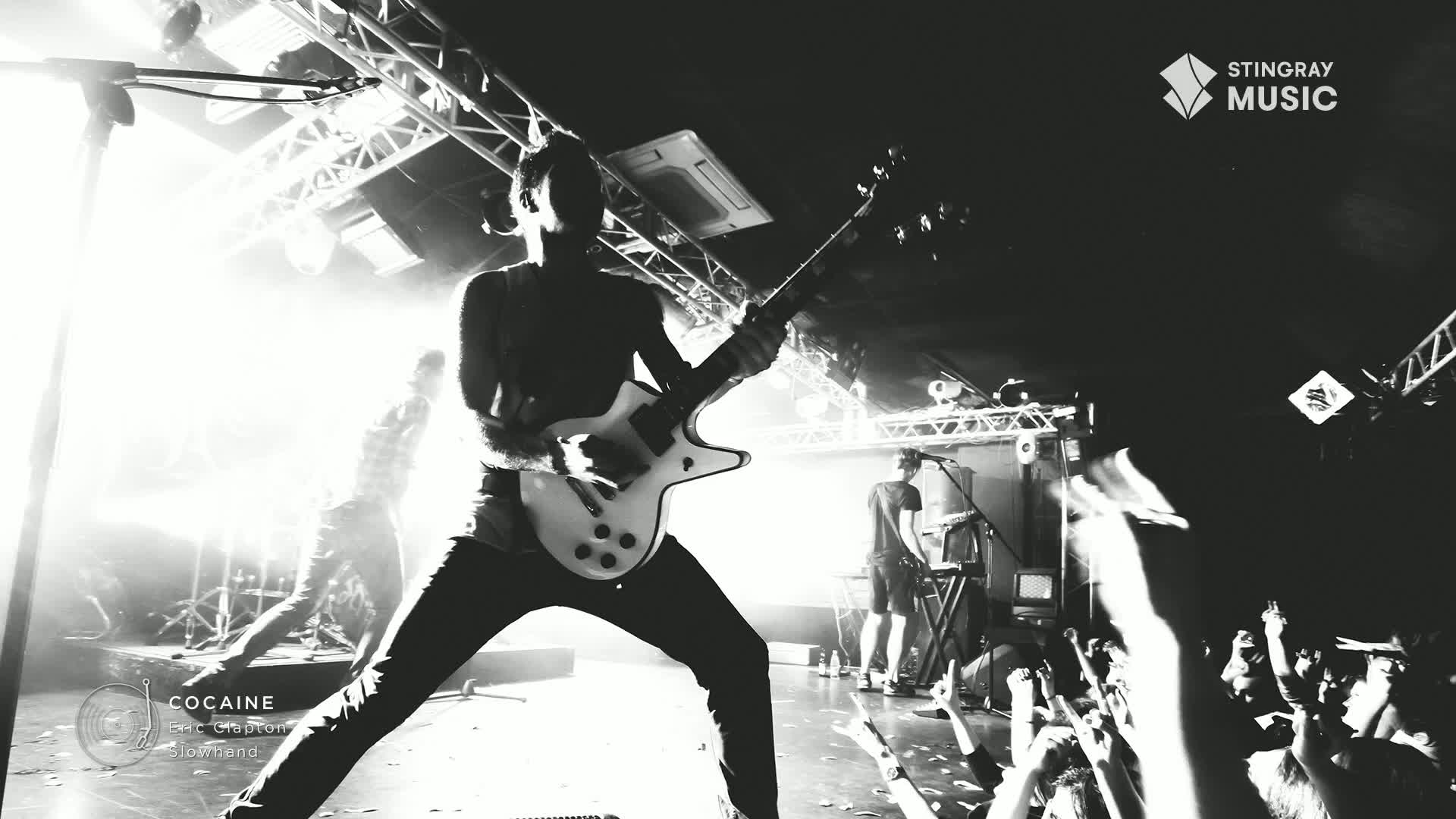 A guitarist, bathed in bright light, is mid-stride, his legs wide as he strums a white electric guitar. The crowd's hands are raised, reaching towards the stage, as the band plays on a Stingray Classic Rock show in Canada.
