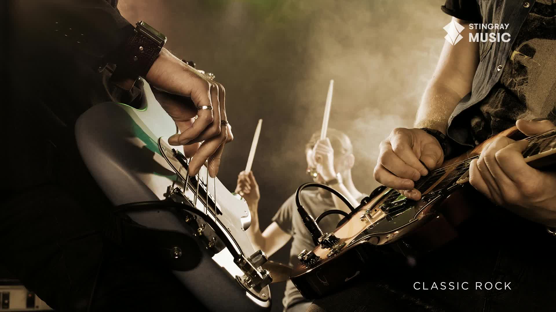 Fingers dance across the fretboard of a bass guitar, while a guitarist strums nearby. Behind them, the drummer's sticks flash in the hazy light, all part of a Stingray Classic Rock performance.
