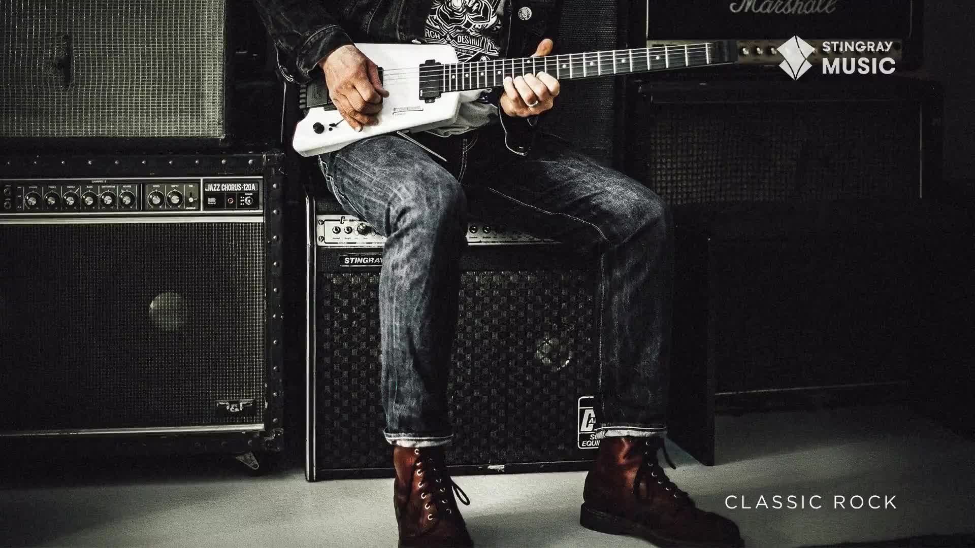 A musician, clad in denim and boots, sits perched atop a speaker stack, fingers dancing across the fretboard of a white guitar. The Stingray Music logo is visible, a nod to the Classic Rock tunes being crafted in this moment.

