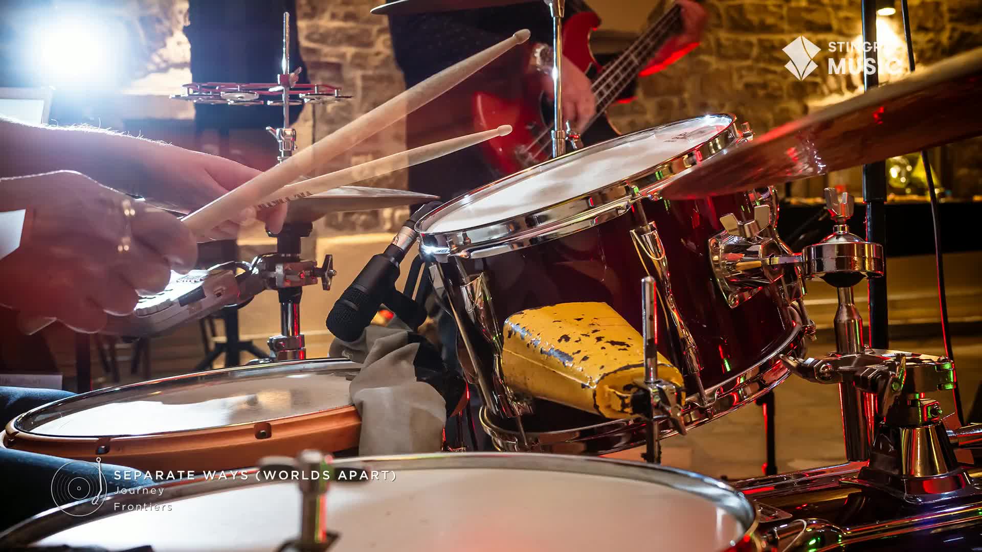 Drumsticks blur as they strike the snare, a driving beat for this classic rock tune. A bass guitar player's hands move over the fretboard in the background.