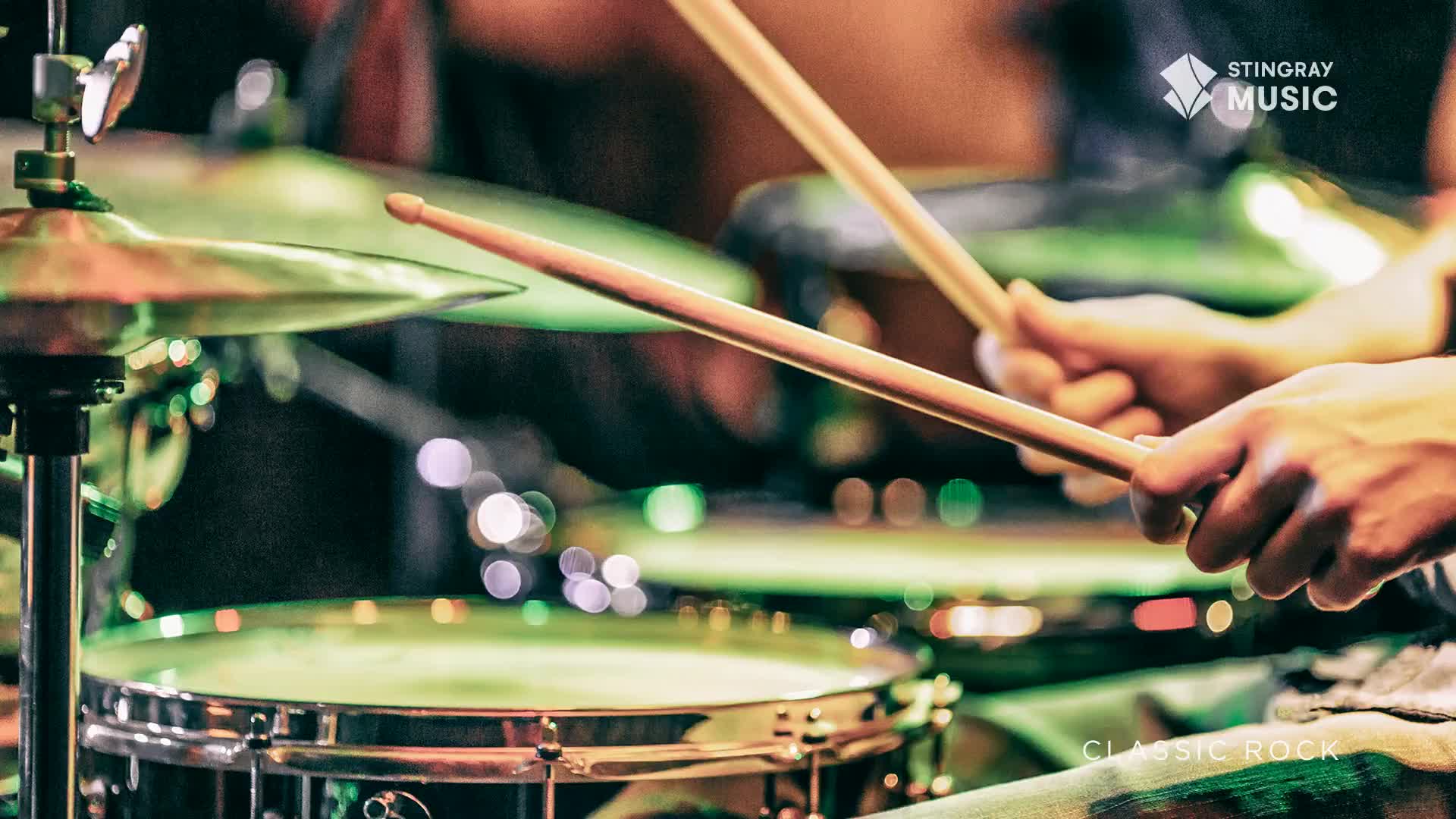 Drumsticks are poised, ready to strike the snare and cymbal. The stage lights cast a green glow on the Stingray Music set, hinting at a classic rock performance.