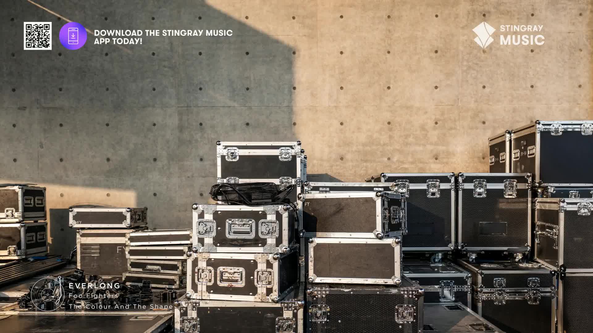 The sun casts long shadows across a wall of stacked, black equipment cases. A black bag rests on top of a case, its straps spilling over the side.