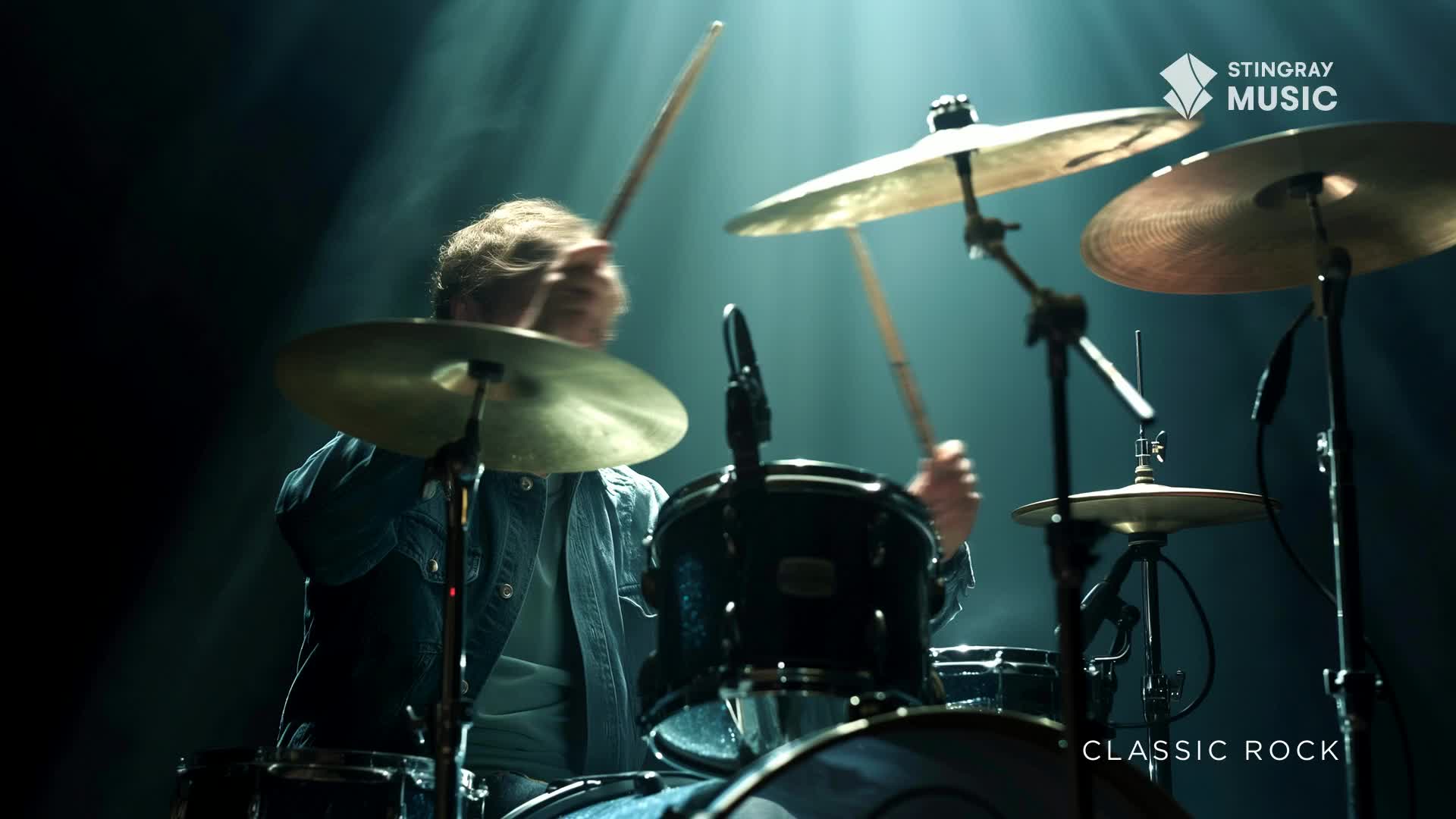 The drummer's hands are a blur as they strike the cymbals and drums, driving the classic rock beat. A spotlight cuts through the darkness, illuminating the Canadian musician and his kit.