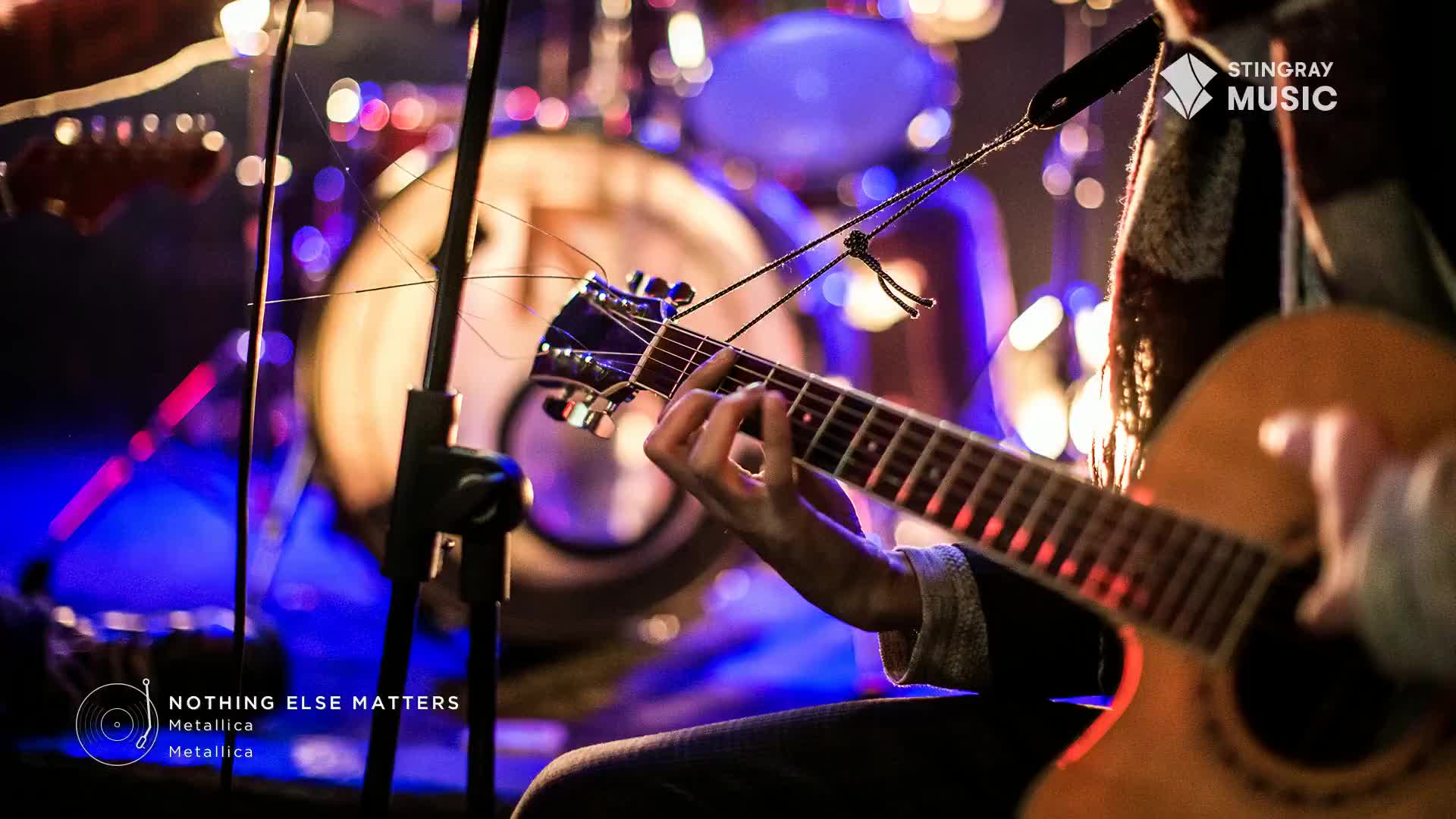 Fingers dance across the fretboard of an acoustic guitar, the warm wood glowing under stage lights. Behind the player, a drum kit waits, bathed in the cool blues and purples of the concert hall.
