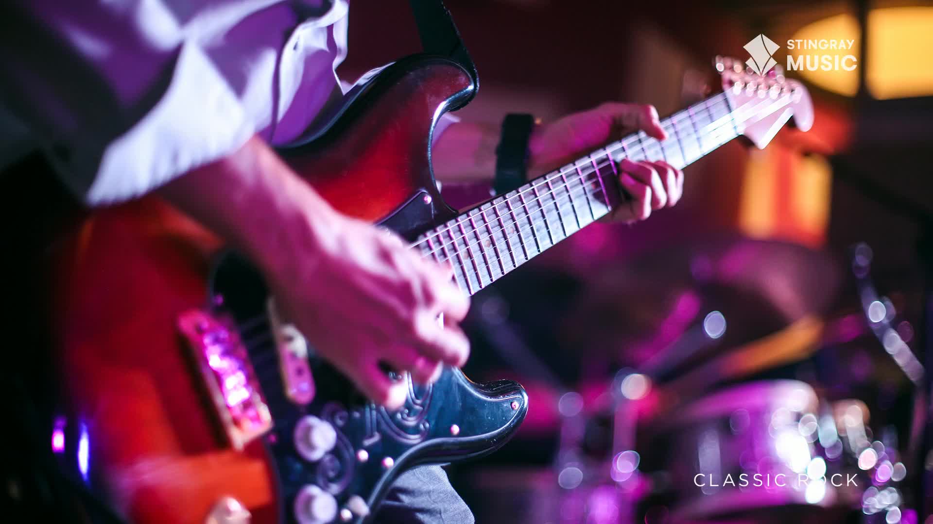 A guitarist's fingers dance across the fretboard of a sunburst electric guitar, bathed in the purple glow of stage lights. Behind him, the drums are a blur of motion, ready for the next classic rock anthem.