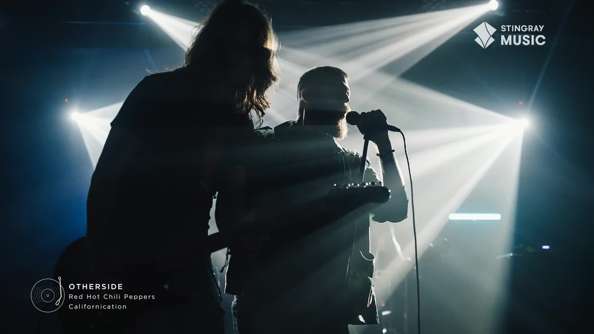 A singer with a beard belts into a microphone, his guitar held loosely at his side. Behind him, another musician with long hair is silhouetted against the stage lights.