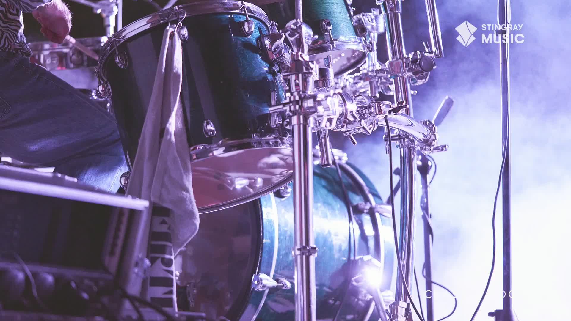 A drummer's hands are a blur as they strike the cymbals and drums, the Stingray Music logo glowing faintly behind the kit. The stage lights cast a purple haze over the scene, hinting at a classic rock performance in Canada.