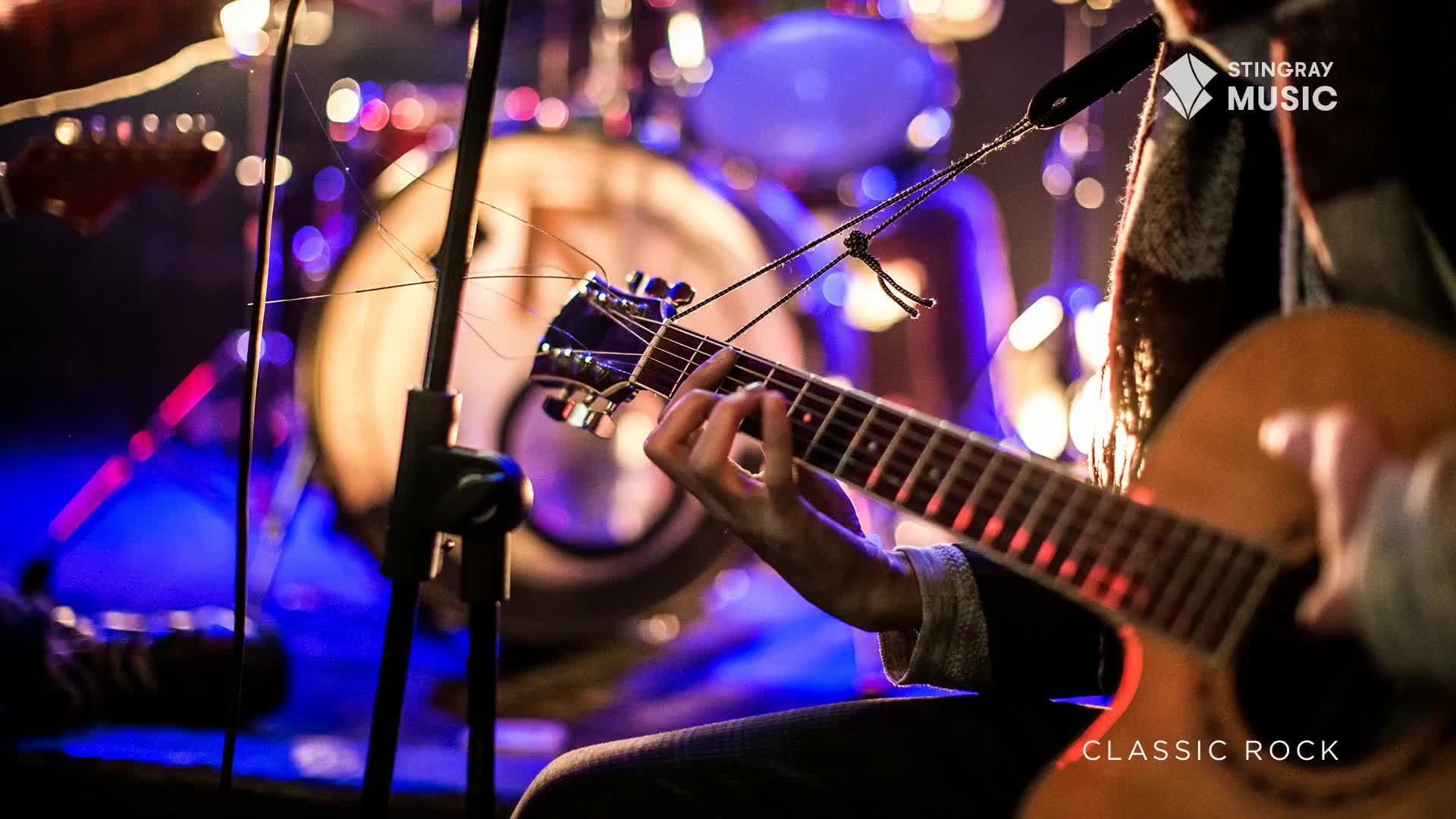 Fingers dance across the fretboard of an acoustic guitar, the warm glow of stage lights catching the wood. Behind, a drummer is a silhouette against the vibrant blues and purples of the stage lighting, ready to lay down a classic rock beat.