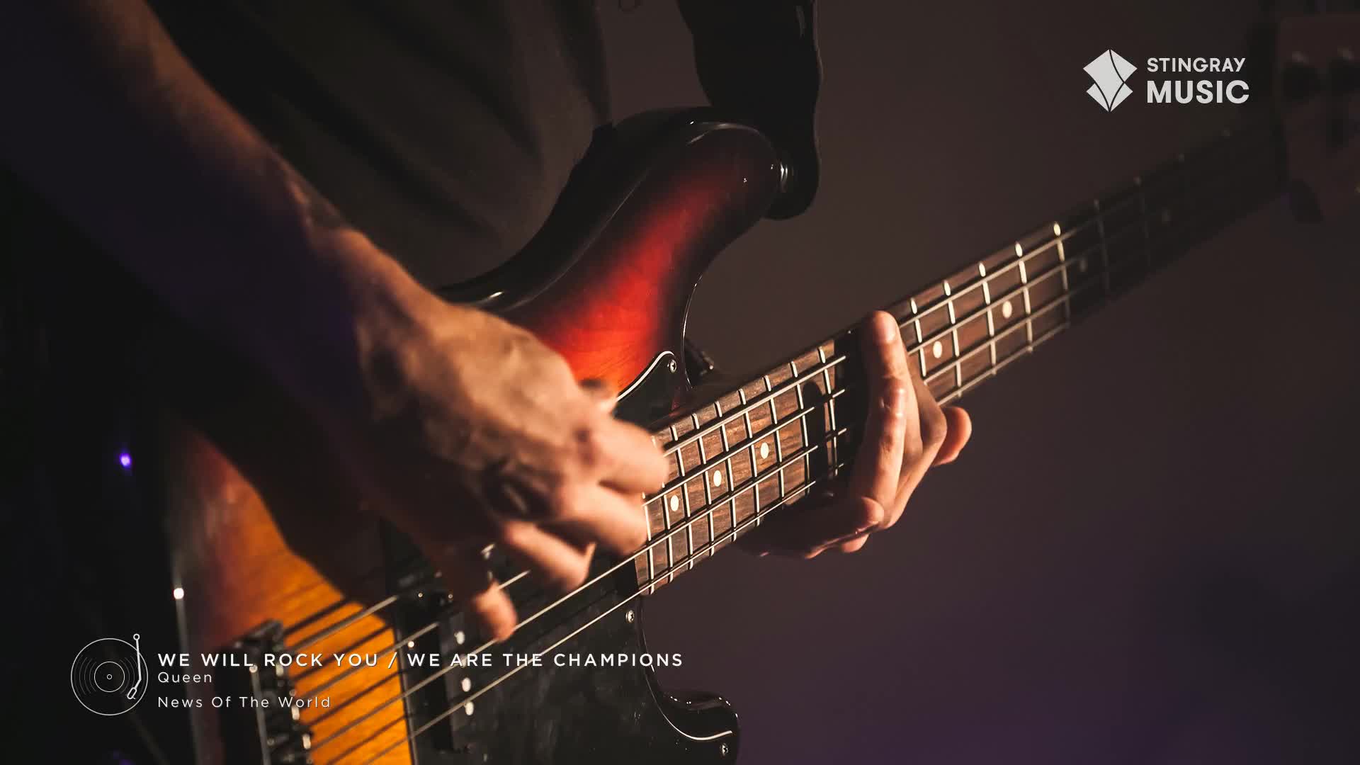 A bass guitar player's hands move across the fretboard, fingers pressing down on the strings. The sunburst finish of the instrument catches the light as the music, a classic rock staple from Queen, fills the air.