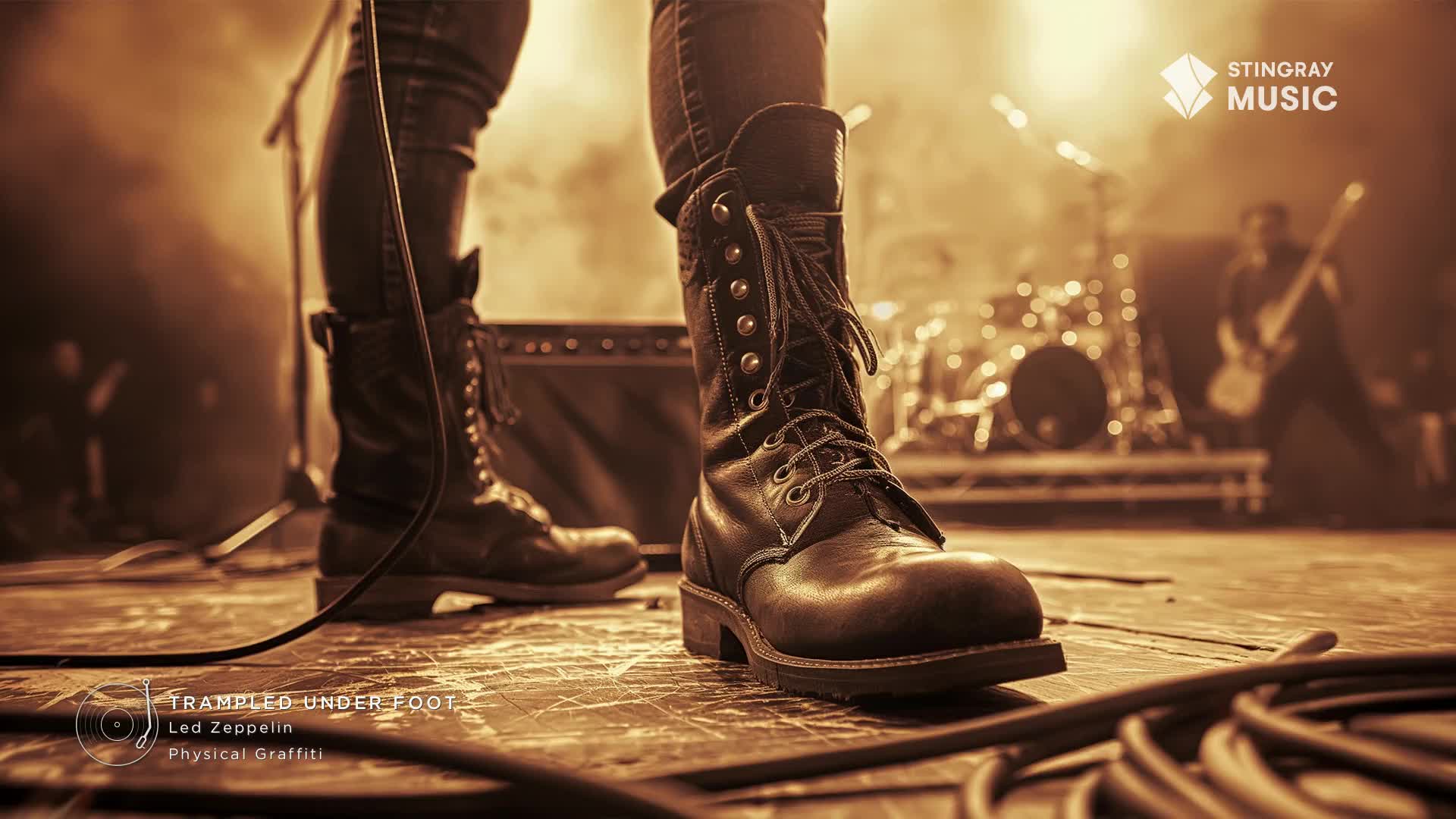 A guitarist moves across the stage, bathed in warm light, while a drummer's kit sits ready behind him. Heavy boots stand planted on the wooden floor, a microphone cable snaking across the stage.