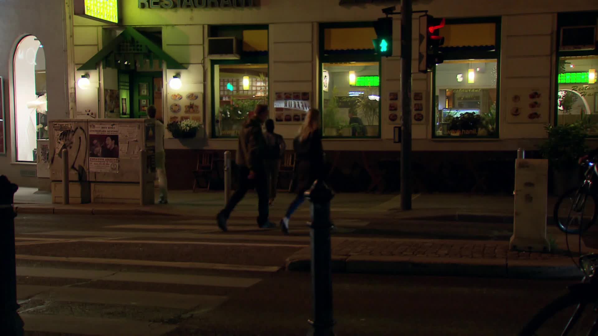 A few people stride across the crosswalk, their silhouettes dark against the illuminated restaurant. The green traffic light shines, and the streetlights cast long shadows on the pavement.
