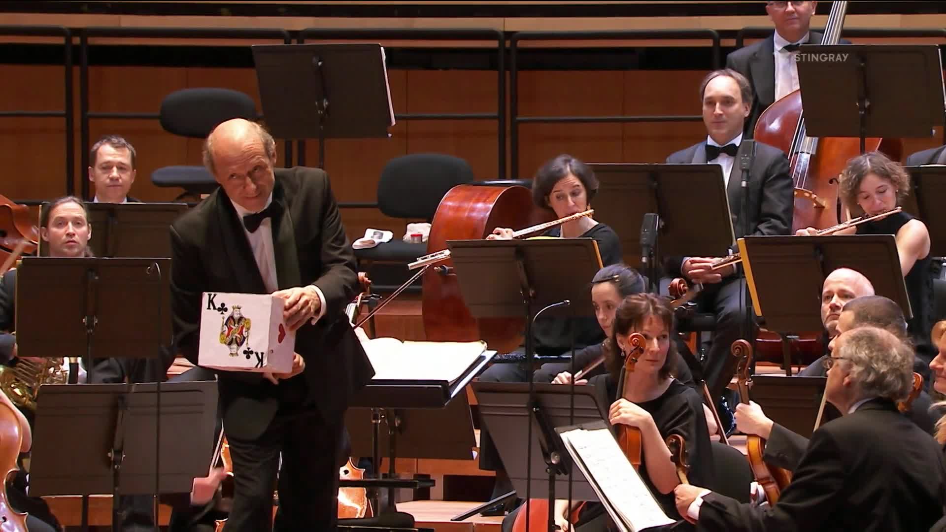 A man in a tuxedo holds a box with a playing card design, facing the orchestra. The musicians, part of a Stingray Classica performance, look on as he presents it.
