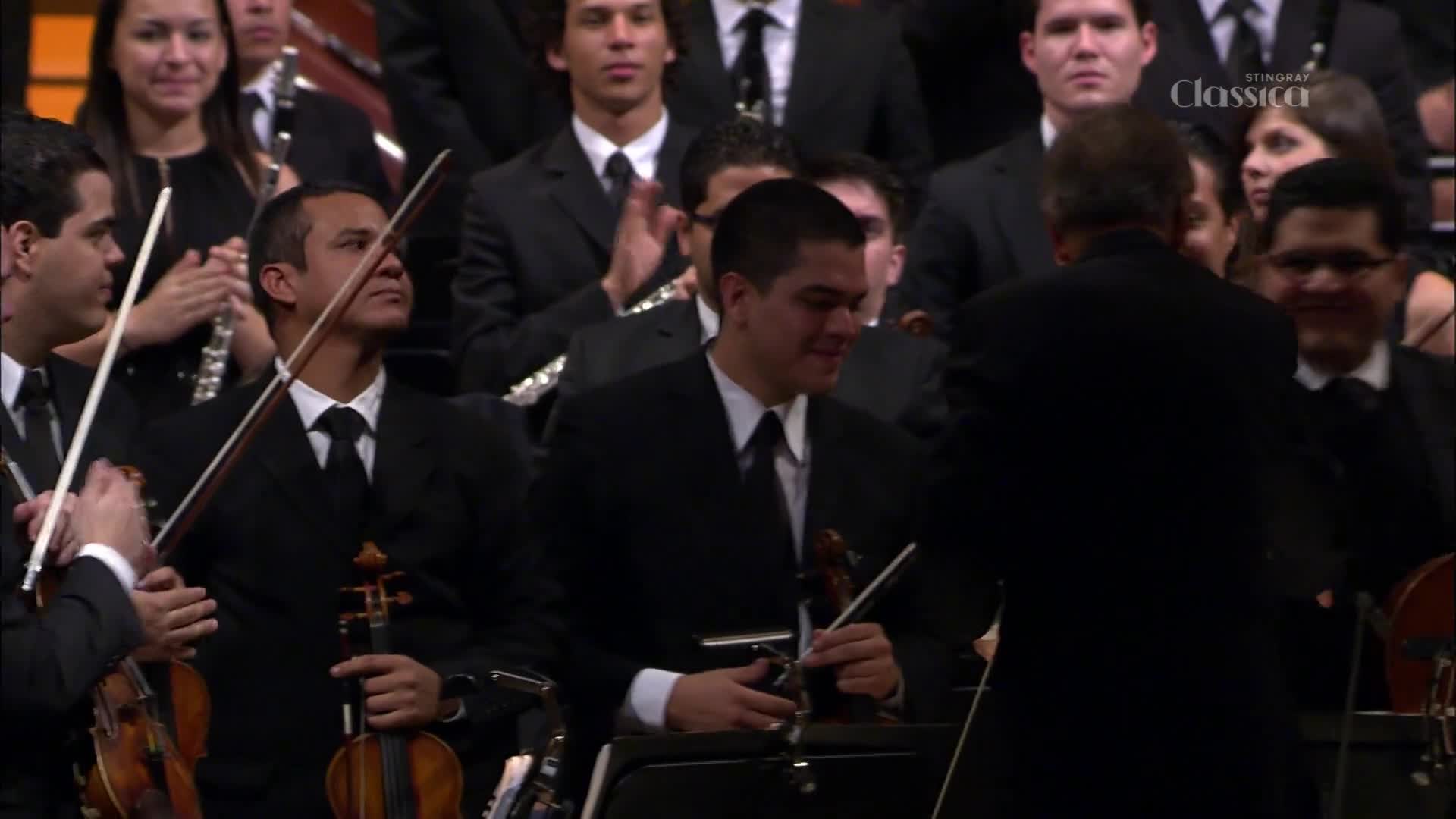 The conductor's back is to me, but I can see the musicians in their black suits and white shirts. Some are holding their violins, while others are clapping, and the Stingray Classica logo is visible.
