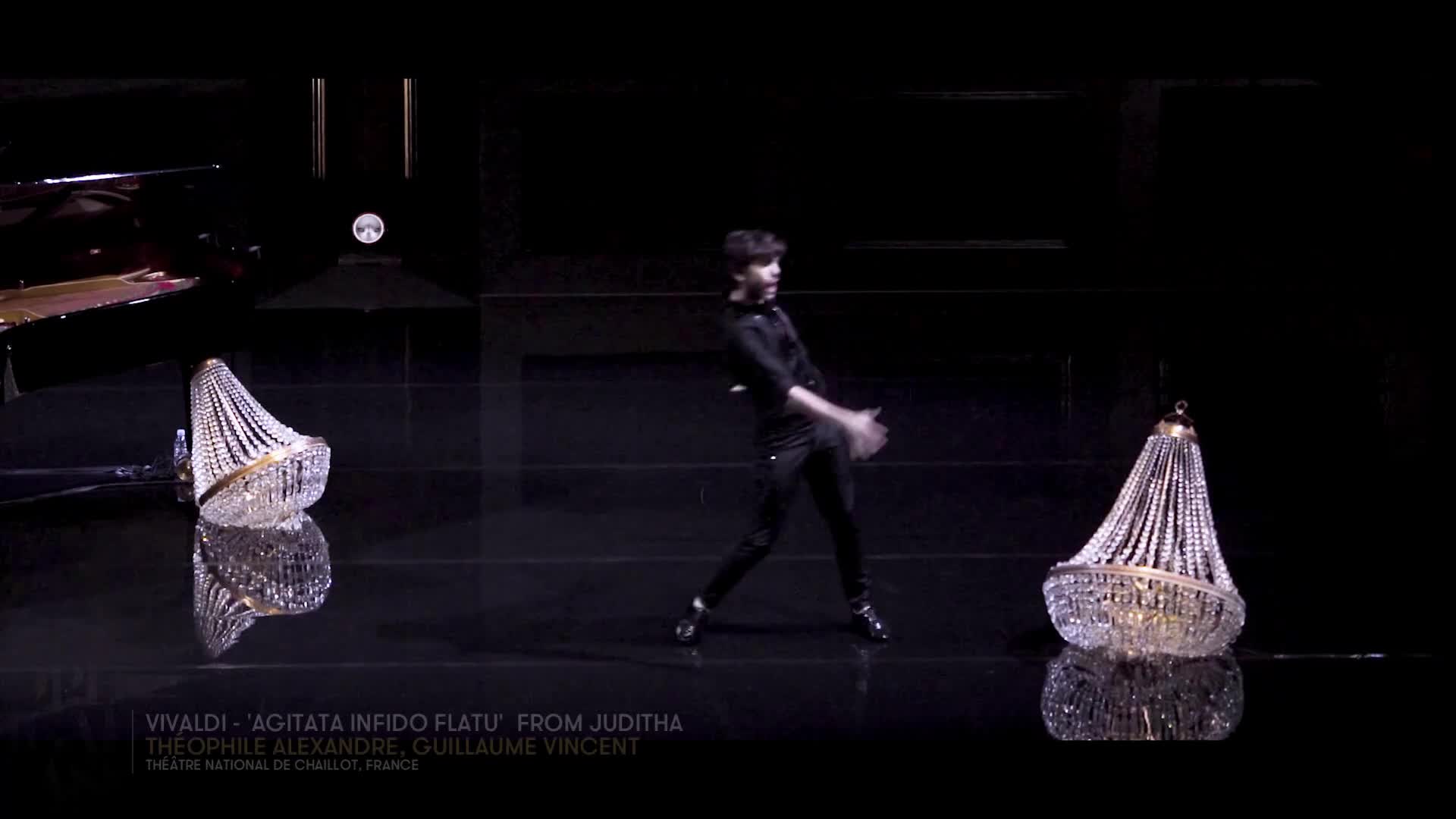 A dancer in black moves across the stage, his arms and legs in motion. Two ornate chandeliers flank him, their crystal beads reflecting the stage lights.
