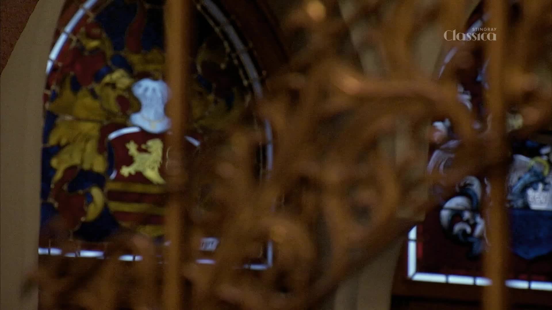 Through a wrought-iron screen, a stained-glass window displays a coat of arms in red, yellow, and blue. The "Stingray Classica" logo is visible in the upper right corner.
