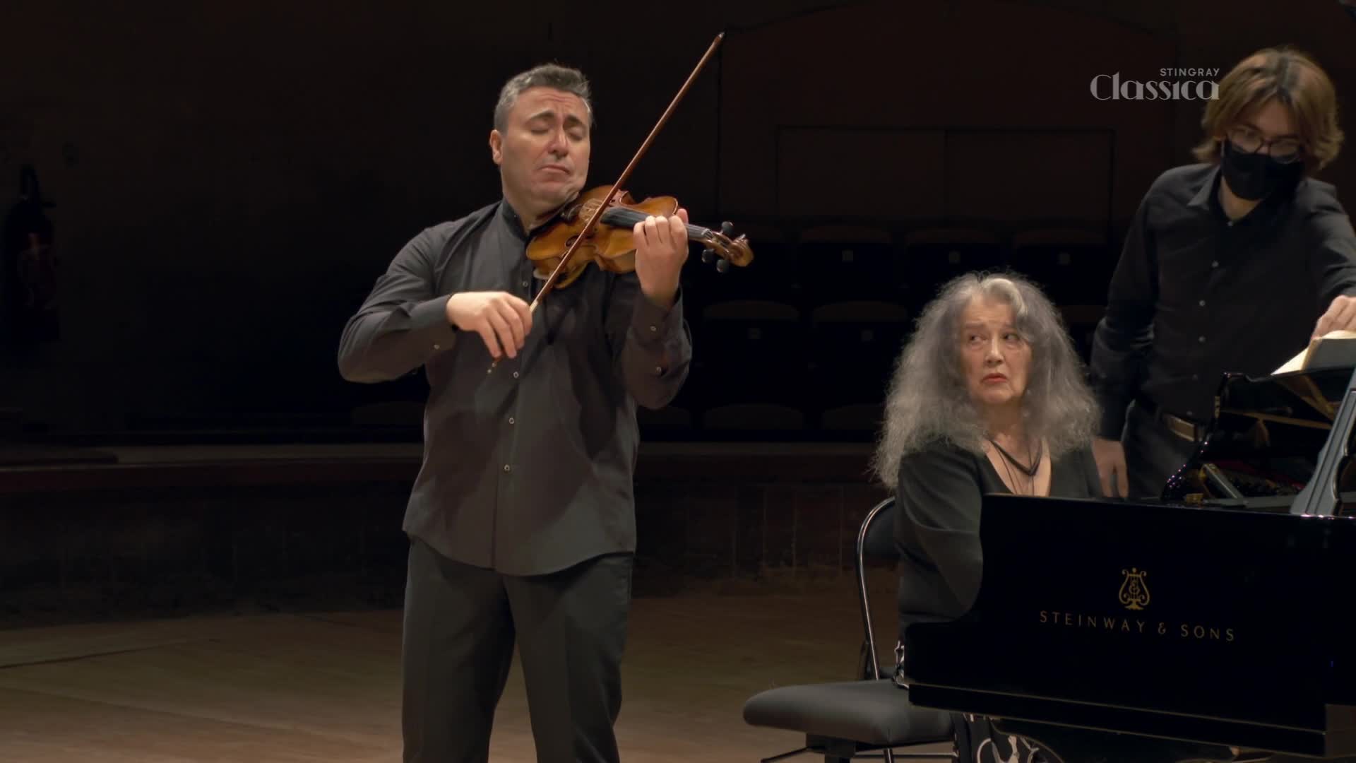 The violinist's bow sweeps across the strings, the sound filling the space. A pianist and a masked assistant are nearby, ready for their cues.
