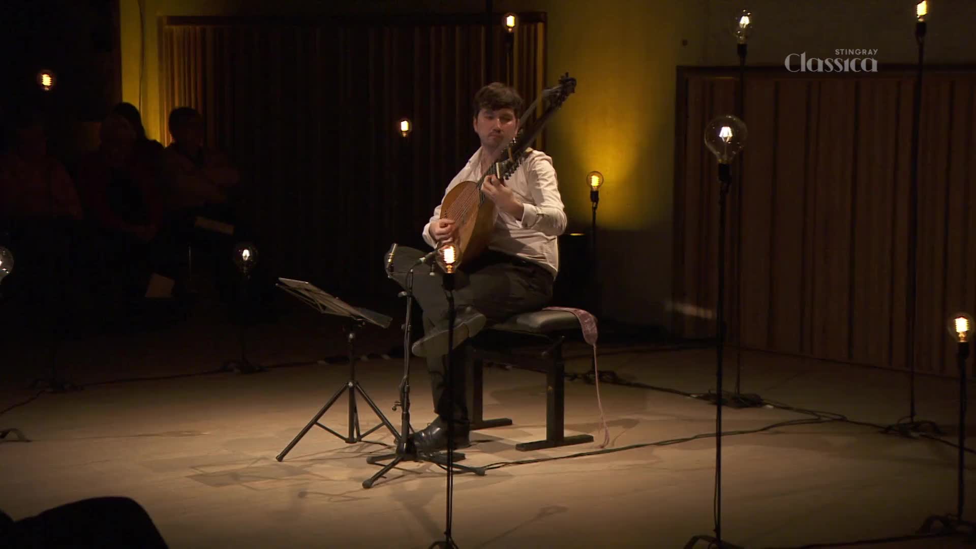 A musician sits on a stool, plucking at a lute-like instrument. The stage is dimly lit, with vintage-style bulbs casting a warm glow around the performer, as if for a Stingray Classica broadcast in Canada.
