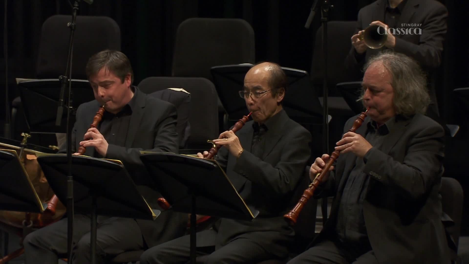Three musicians, each in a black suit, are intently playing their instruments. The woodwind instruments, with their dark wooden bodies, are held close to their faces as they blow.
