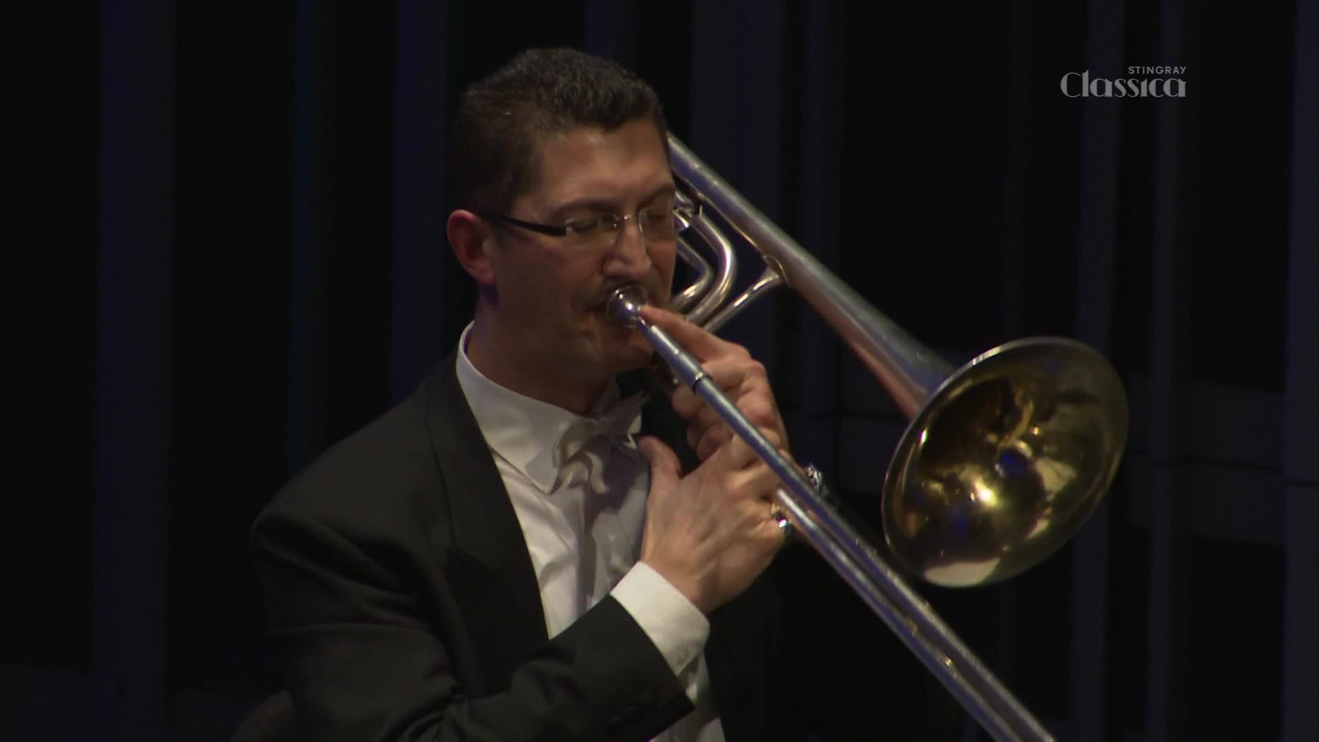 A man in a tuxedo is playing a trombone, the silver slide moving back and forth as he blows. The Stingray Classica logo is visible in the corner.
