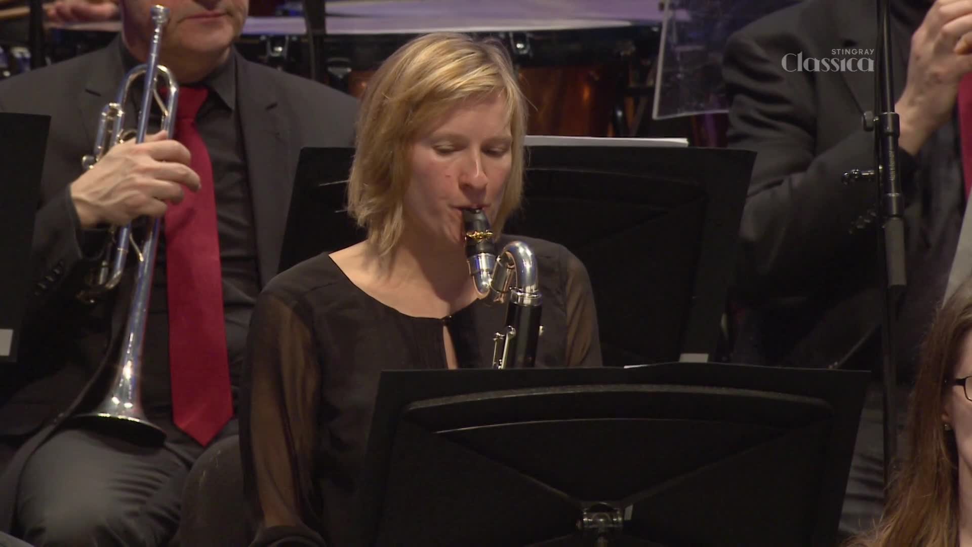 A woman in a black dress is playing a clarinet, her eyes closed in concentration.  Behind her, a man in a suit and red tie holds a trumpet, part of a larger orchestra on Stingray Classica, Canada.
