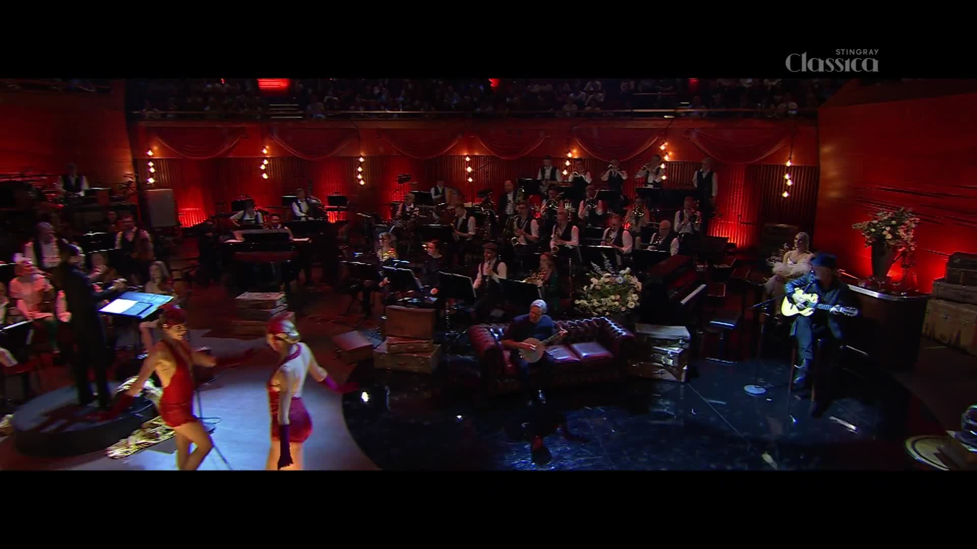 A stage is set for a performance, with an orchestra seated in the background. Two dancers in red costumes move in front of the orchestra as a man with a guitar sings.
