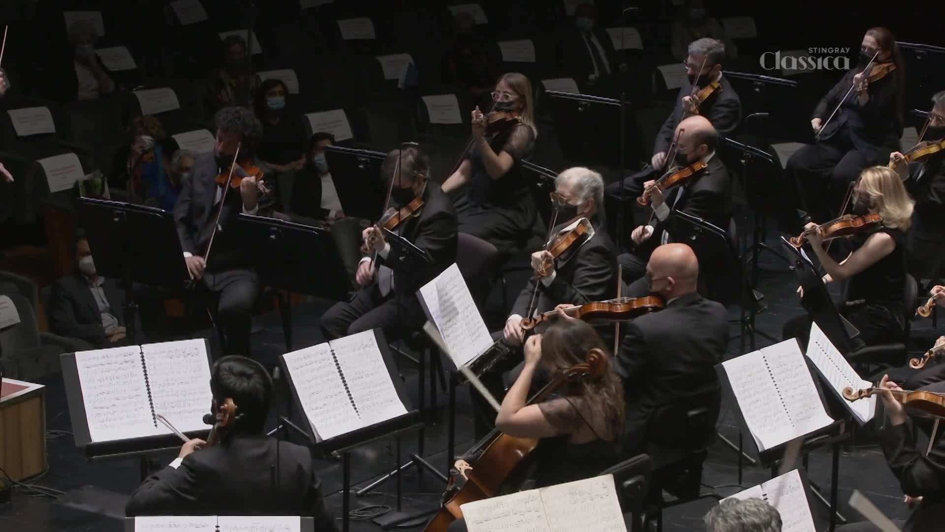 The musicians, masked, are bent over their instruments, bowing in unison. The conductor's arm is raised, poised to bring down the baton.
