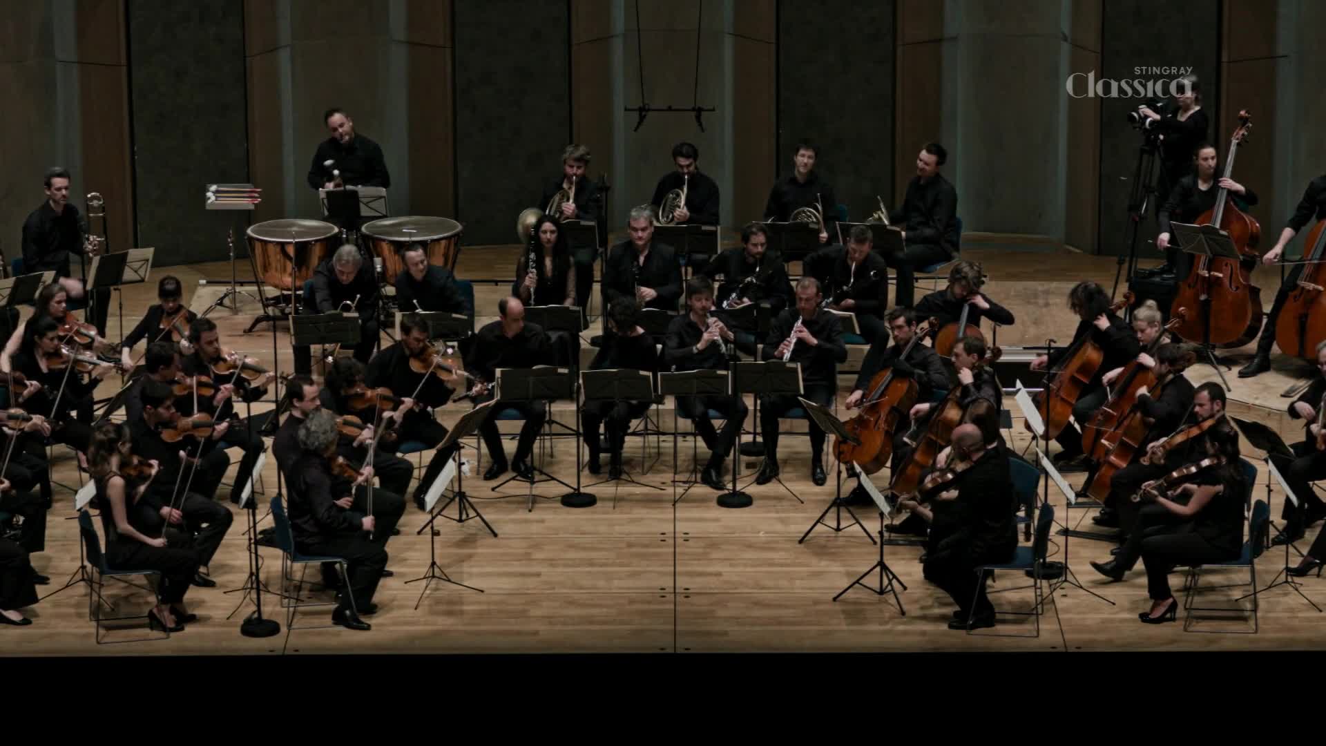 An orchestra, likely in Canada, is performing on stage, the musicians focused on their sheet music.  The Stingray Classica logo is visible, and the lighting is focused on the musicians.
