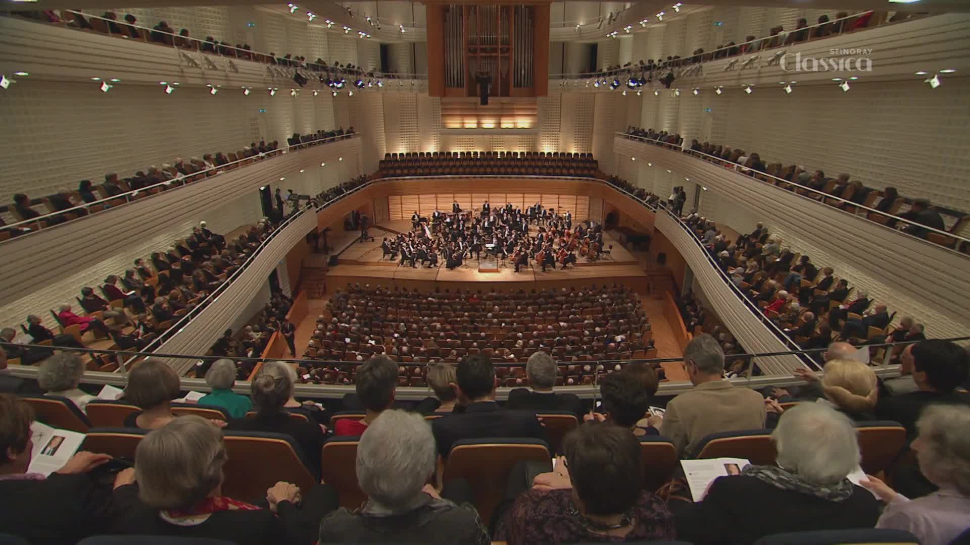The orchestra is poised, instruments glinting under the lights as they prepare to play. A full house of people in formal attire sit quietly, anticipating the performance broadcast on Stingray Classica.
