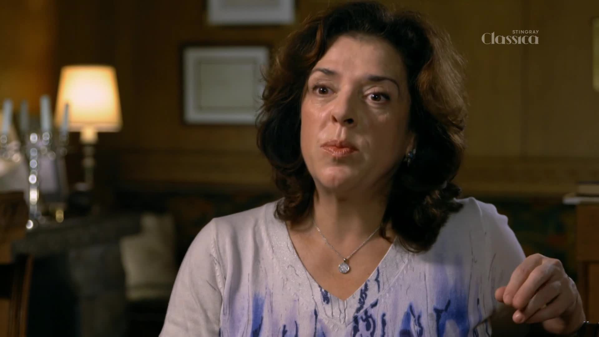 A woman with dark, curly hair is speaking, her lips slightly parted as she pauses. The Stingray Classica logo is visible, and a warm lamp glows in the background.
