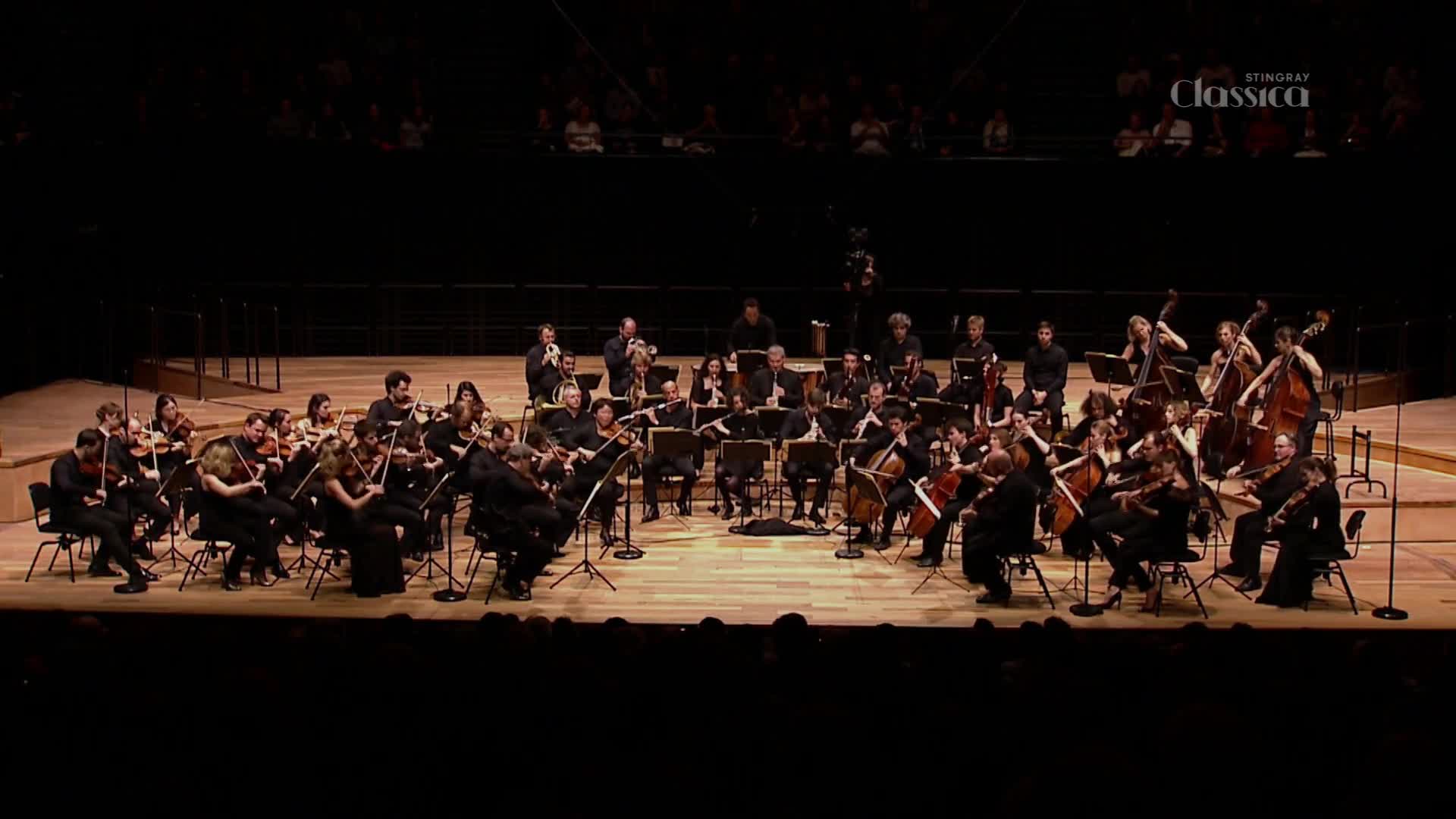 The orchestra, dressed in black, is poised, bows raised, ready to play. The Stingray Classica logo sits in the upper right corner, a subtle reminder of the broadcast.
