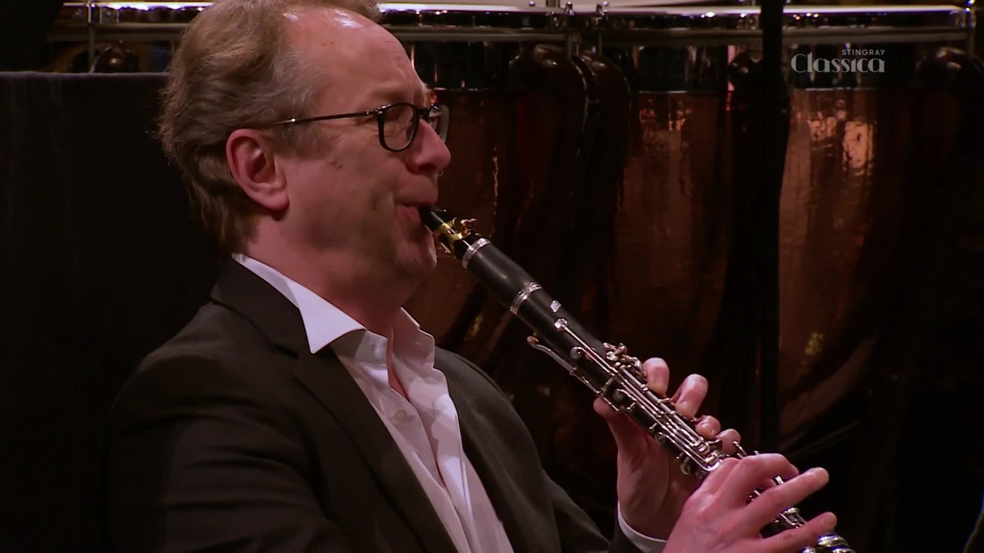 A man in a suit is playing a clarinet, his cheeks puffed out as he blows into the mouthpiece. Behind him, the Stingray Classica logo is visible, and the soft glow of the stage lights reflects off the instruments.
