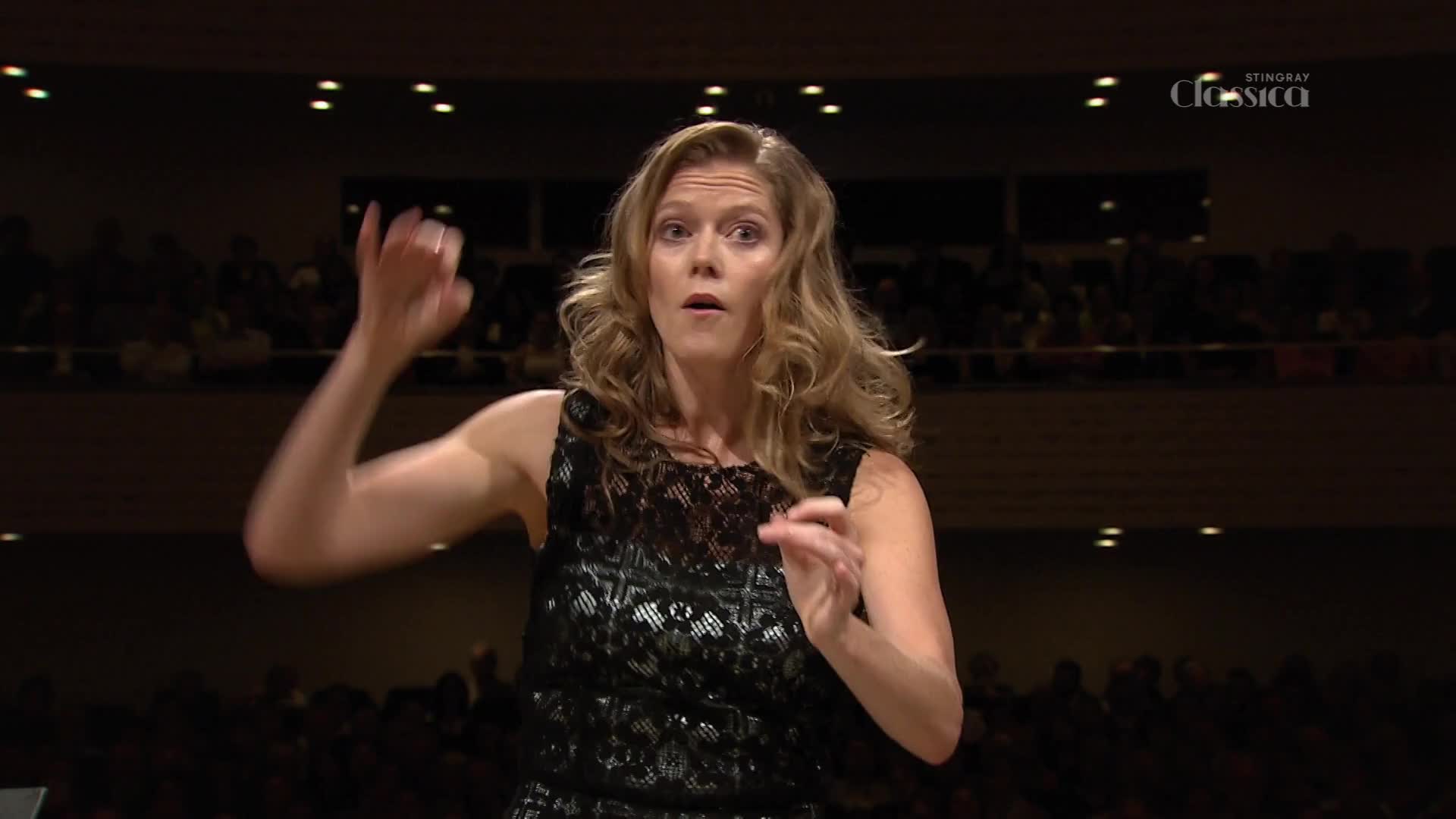The conductor's arm rises, her hand pointing upwards as she guides the orchestra. Her expression is focused, her mouth slightly open as she leads the performance on Stingray Classica.
