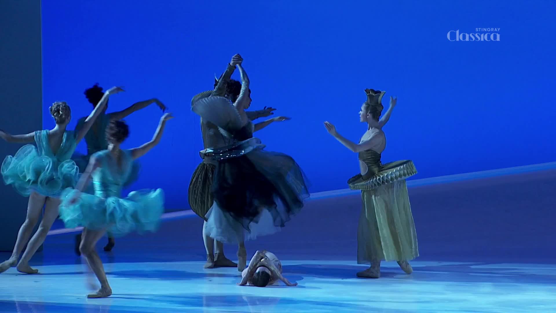 Dancers in turquoise tutus leap and turn, their arms gracefully arched. A figure in a large, dark costume stands with arms raised, while another lies curled on the floor.
