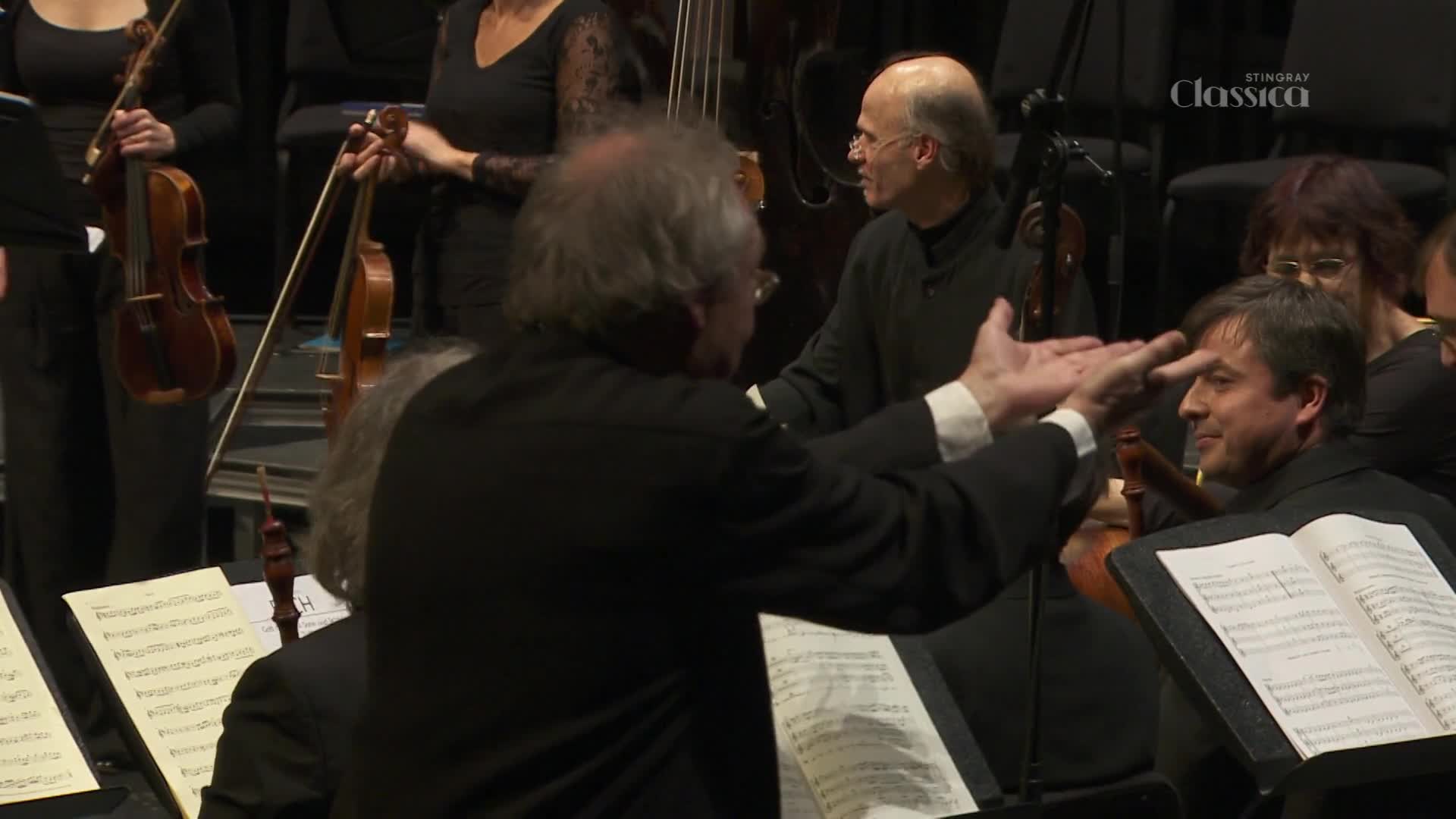 The conductor gestures expansively toward the musicians, his hands open and palms up. A violinist smiles, looking toward the conductor as the orchestra prepares to play.
