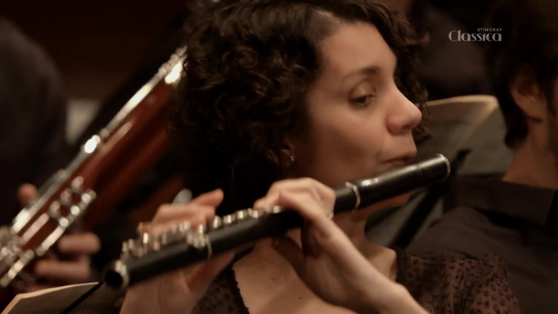 A woman with curly hair is playing a black flute, her fingers dancing over the keys.  Behind her, a bassoon rests, its wooden body partially visible in the dim light of what appears to be a Canadian concert hall, as seen on Stingray Classica.
