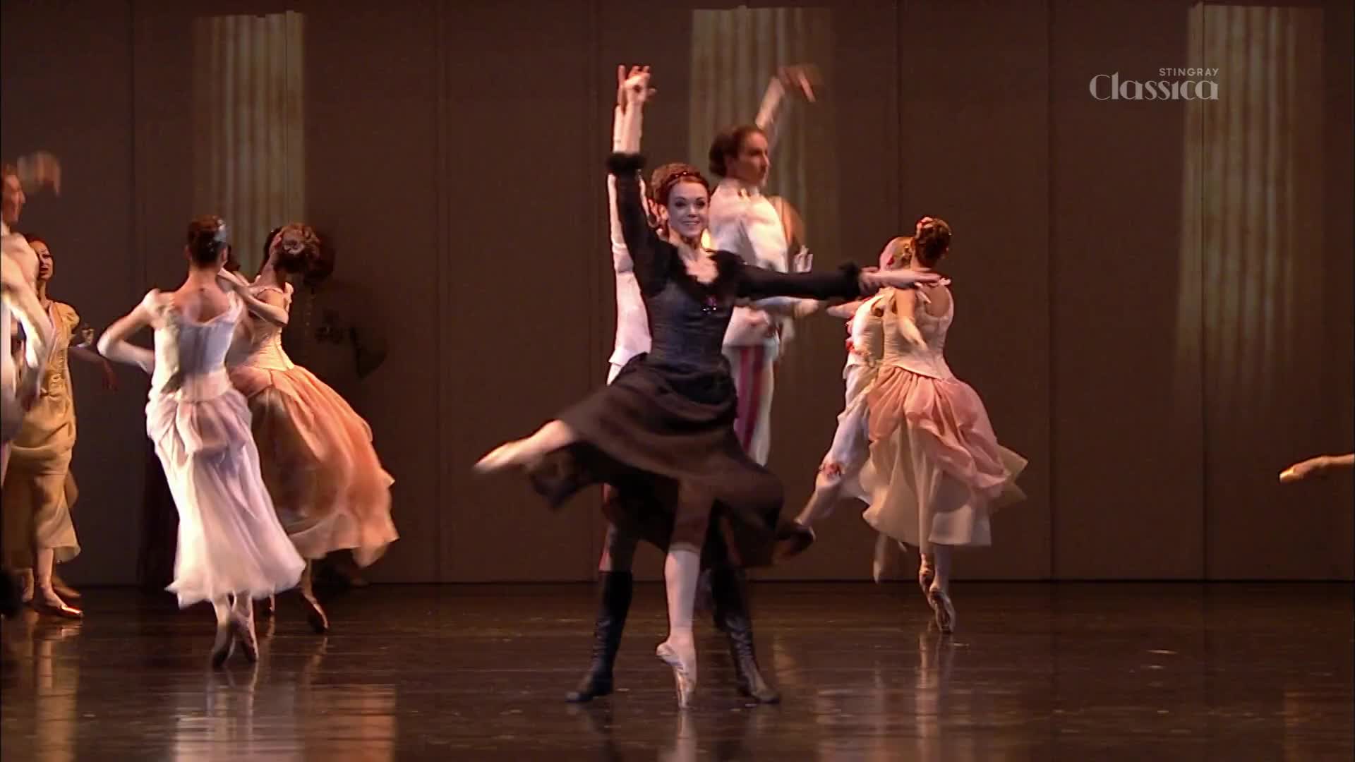 A woman in a black dress is leaping, her arms raised, while a man supports her from behind. Other dancers in pastel gowns and white shirts are scattered across the stage.
