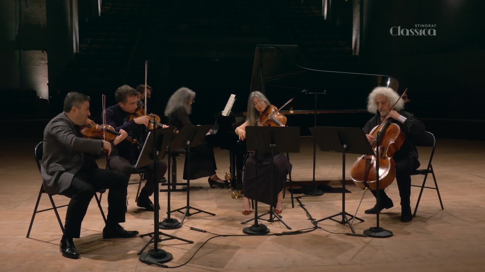 A string ensemble performs on a stage, illuminated by soft lighting. The musicians, including a cellist with white hair, bow their instruments, their movements synchronized.
