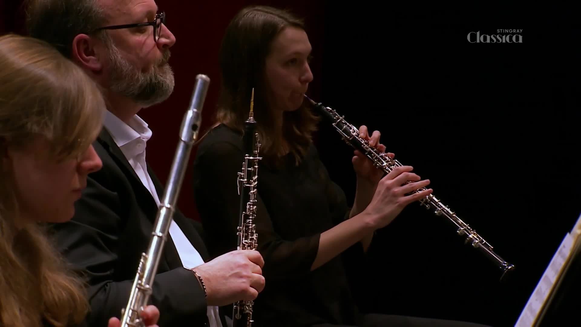 An oboist is blowing into the instrument, her fingers dancing over the keys. Beside her, a flutist and another musician with a flute are focused on their music.
