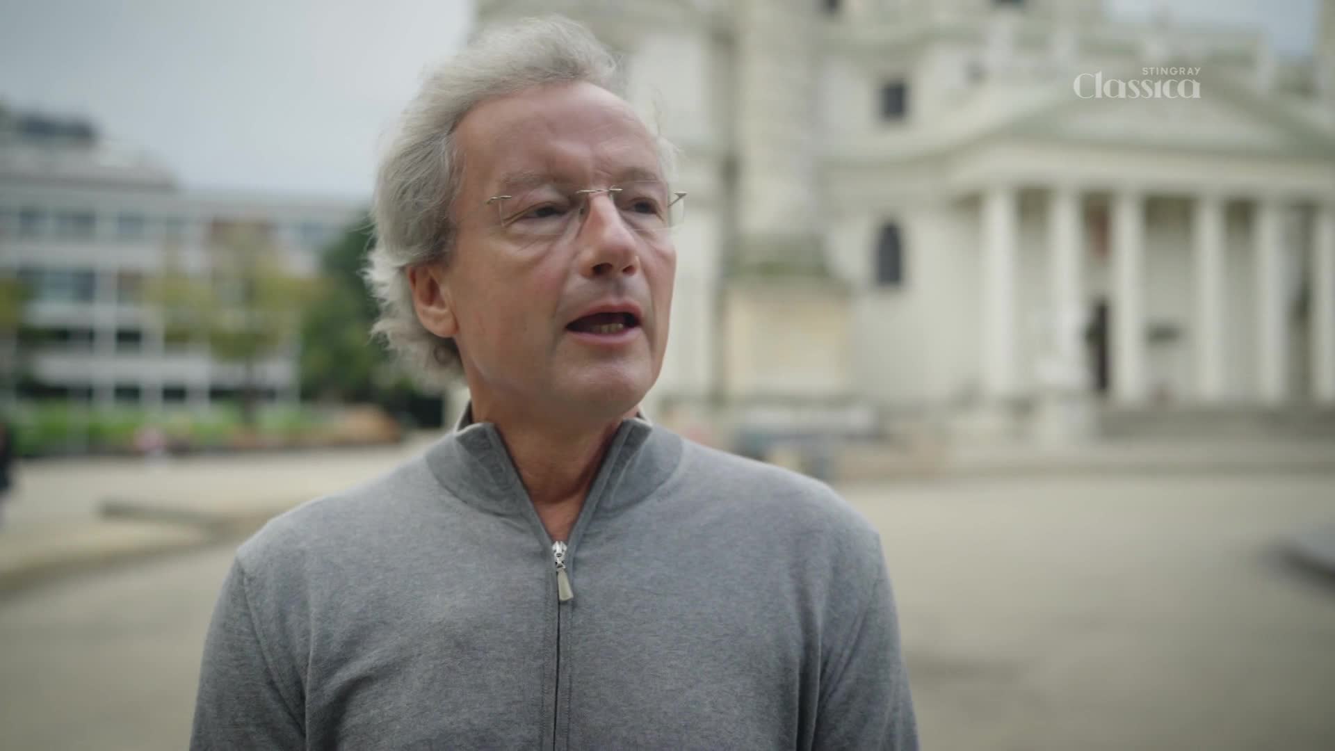 A man in a grey sweater speaks, his mouth open as if mid-sentence. Behind him, a large building with columns and the Stingray Classica logo are visible.
