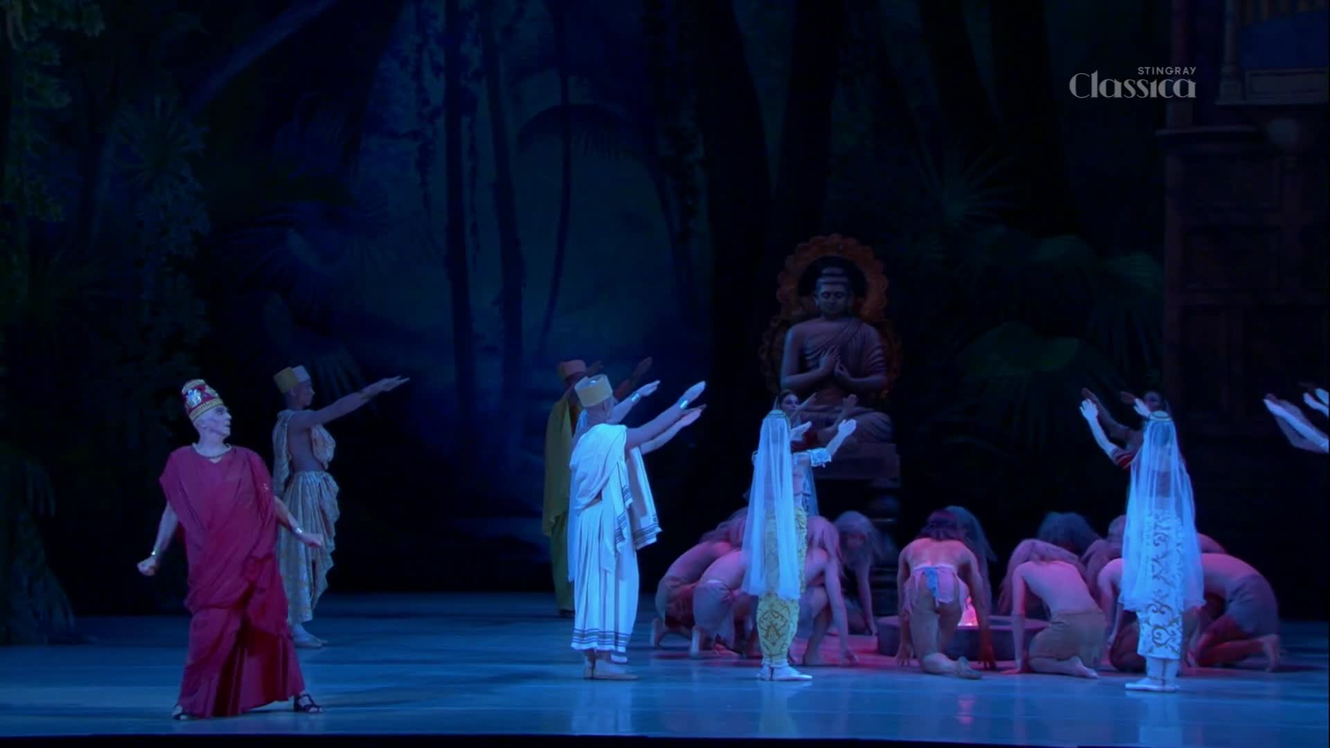 Dancers in flowing robes gesture toward a seated Buddha statue, their arms raised in reverence. The stage, lit in blues and greens, sets the scene for this performance, broadcast by Stingray Classica from Canada.
