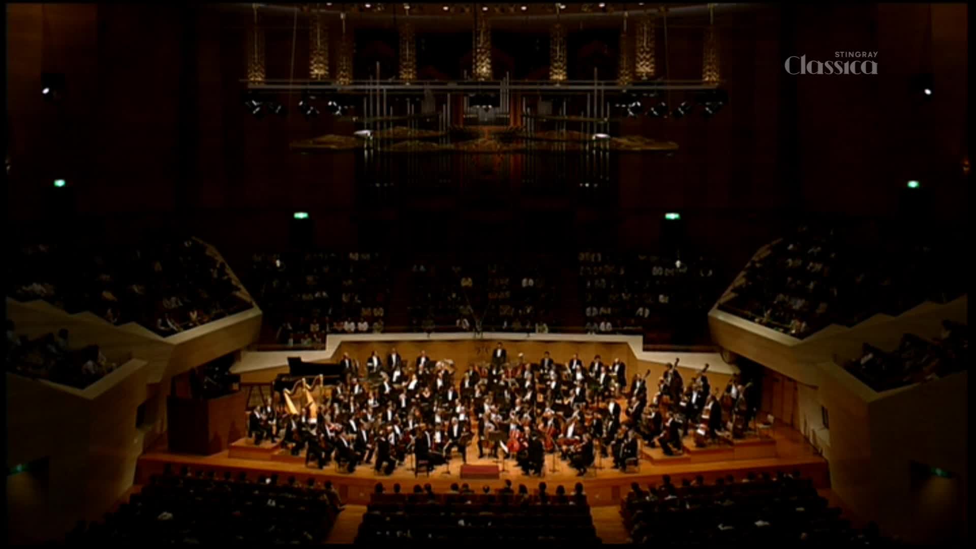 An orchestra, dressed in black tie, is arranged on a stage bathed in warm light. Above, the Stingray Classica logo is visible, and the audience is seated in the darkened hall.
