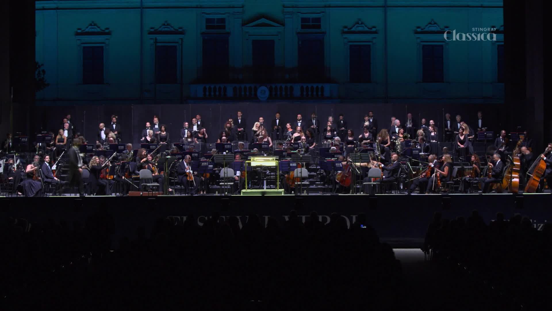An orchestra, bathed in stage light, is tuning their instruments as the conductor walks to his podium.  The backdrop shows a building, and the Stingray Classica logo is visible in the upper right.
