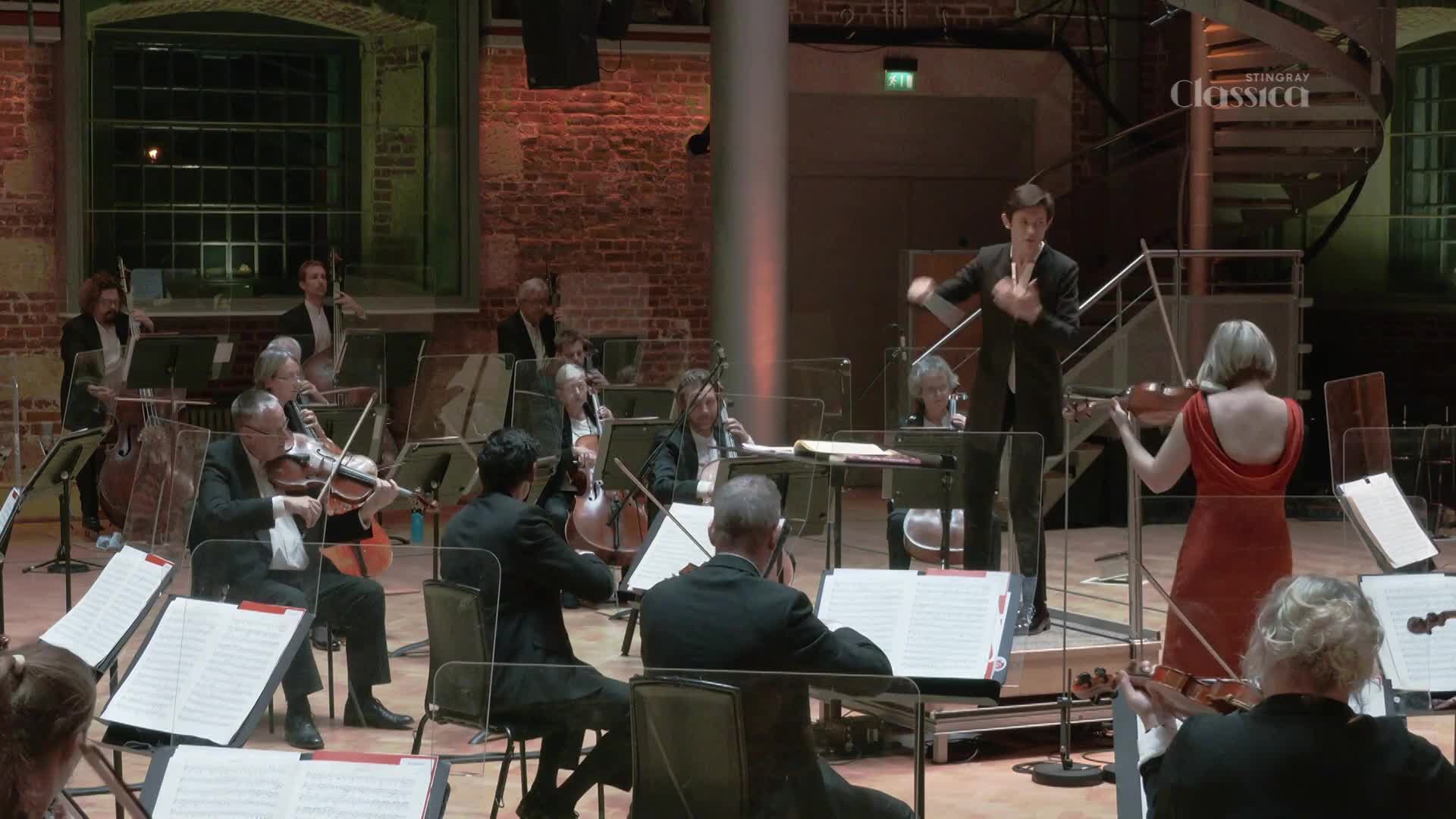 The conductor's arms are raised, guiding the orchestra with broad gestures. A violinist in a red dress stands to the side, her instrument poised.
