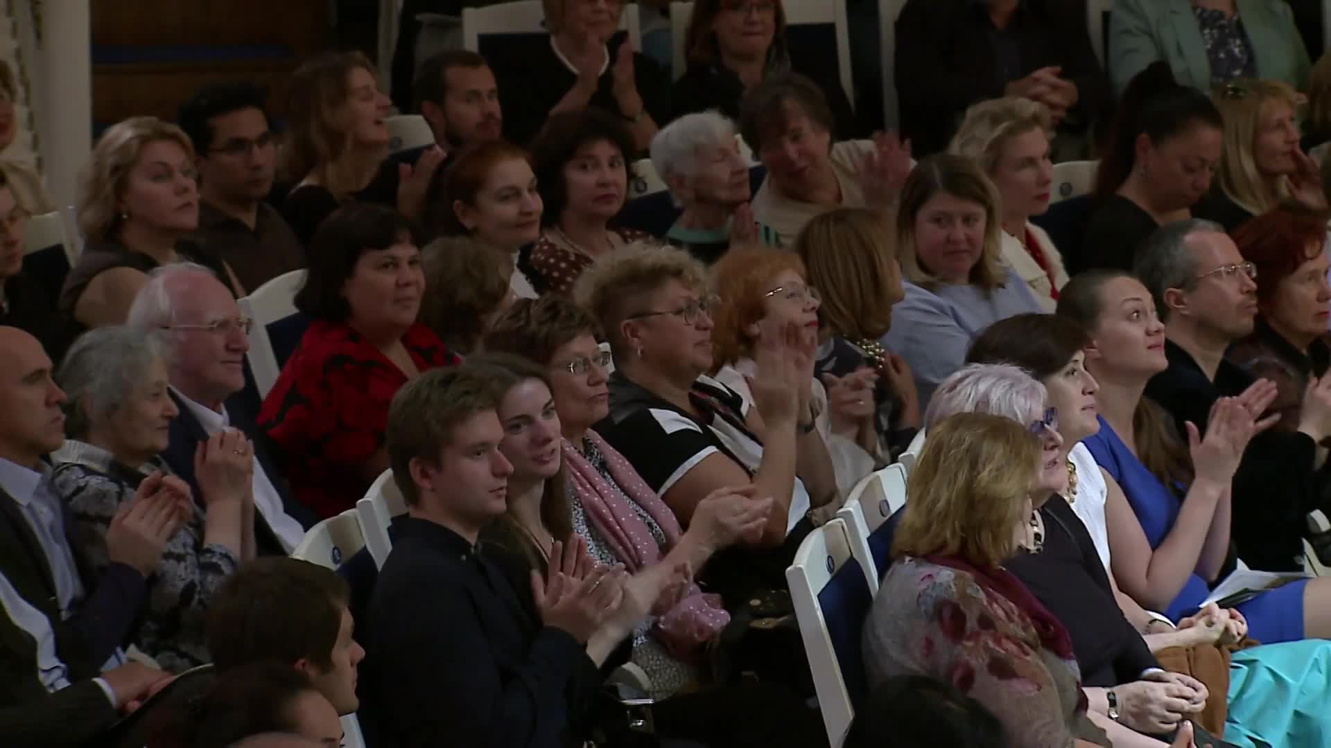 The audience is clapping, their hands rising and falling in unison. A sea of faces, young and old, are turned toward the stage.
