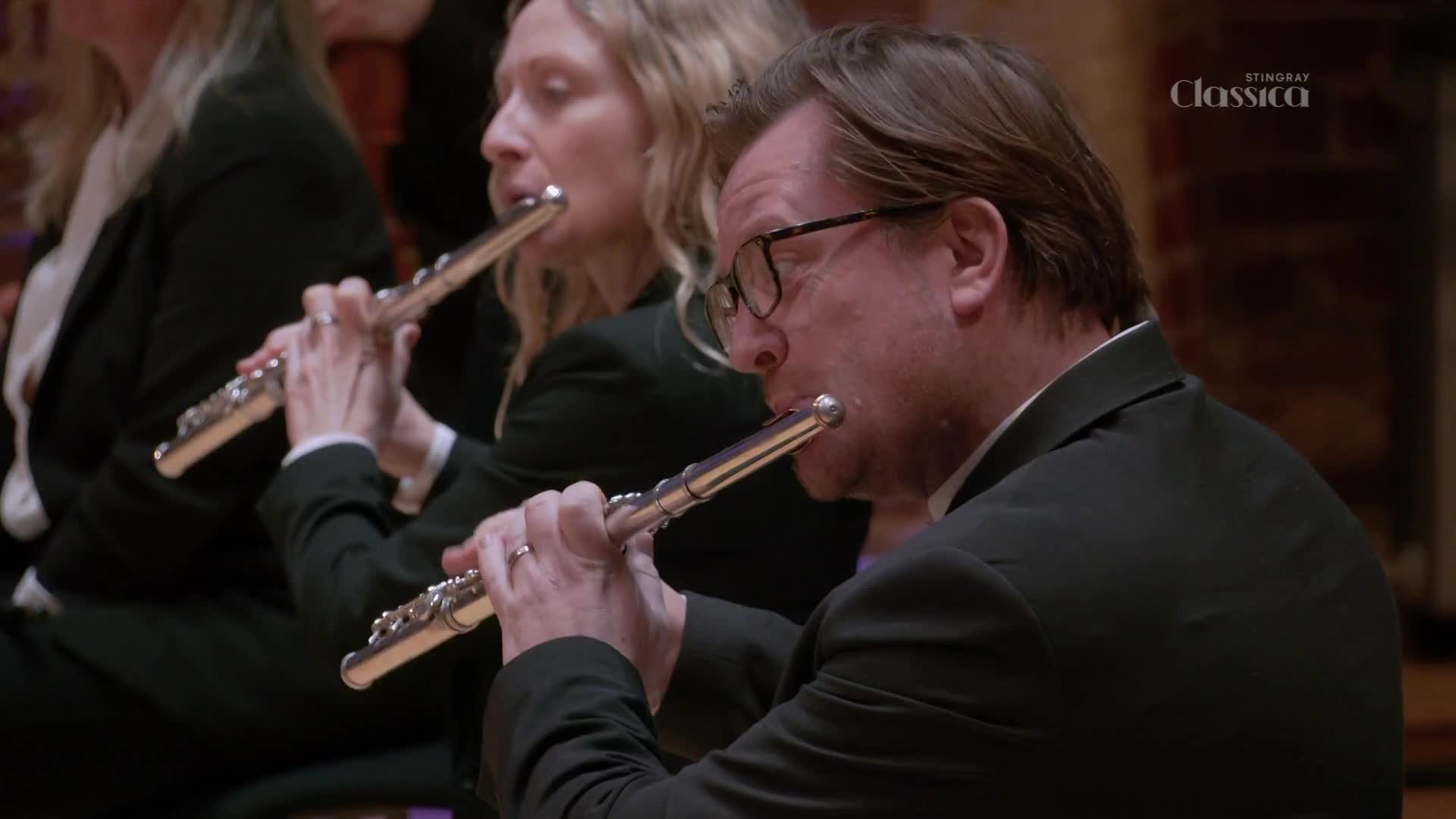 The flutists, dressed in black suits, are focused on their instruments. They blow into the silver flutes, fingers dancing over the keys as they play.
