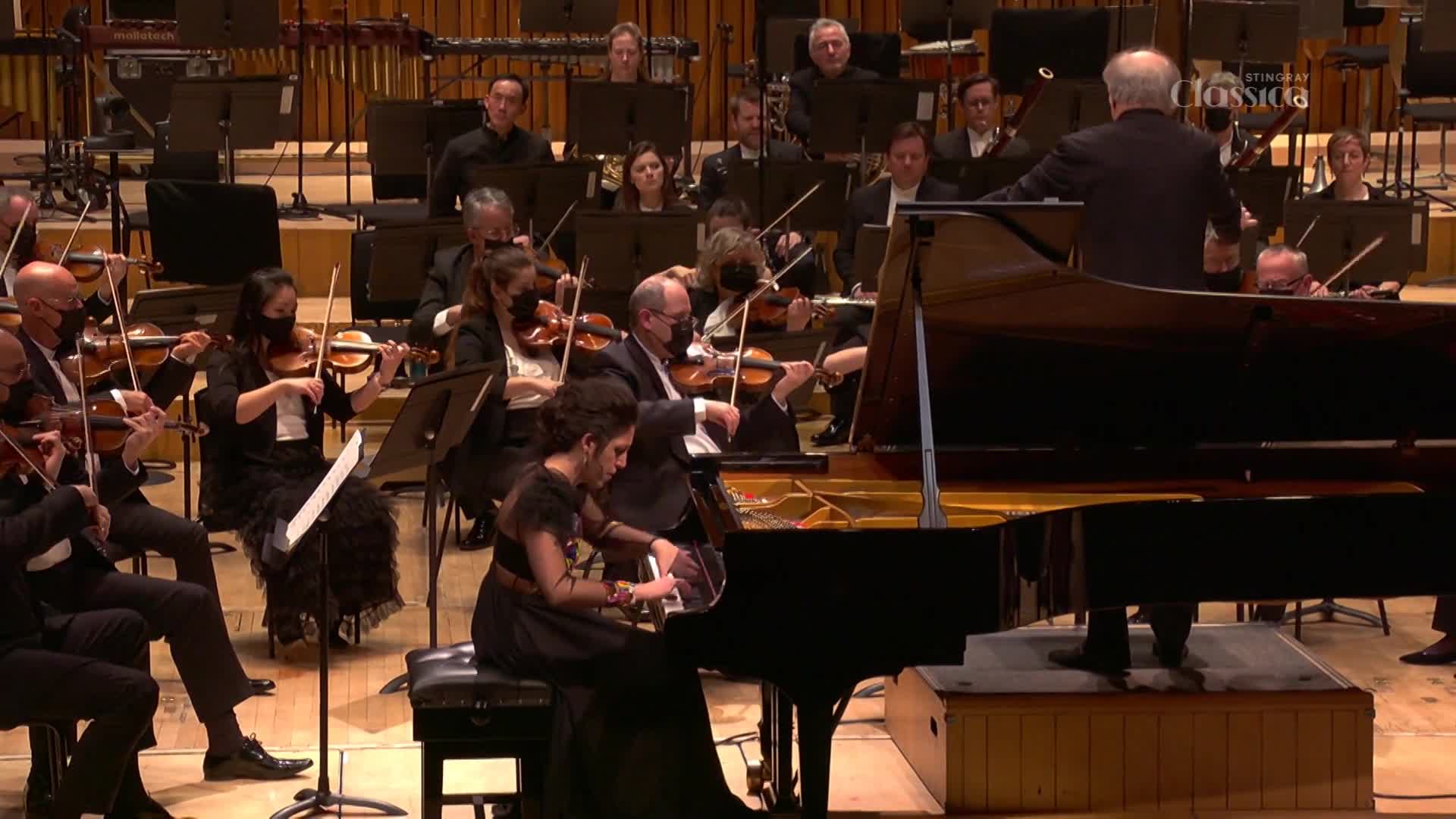 The conductor's arms are raised, leading the orchestra. A pianist's fingers dance across the keys of a grand piano, as the musicians play.
